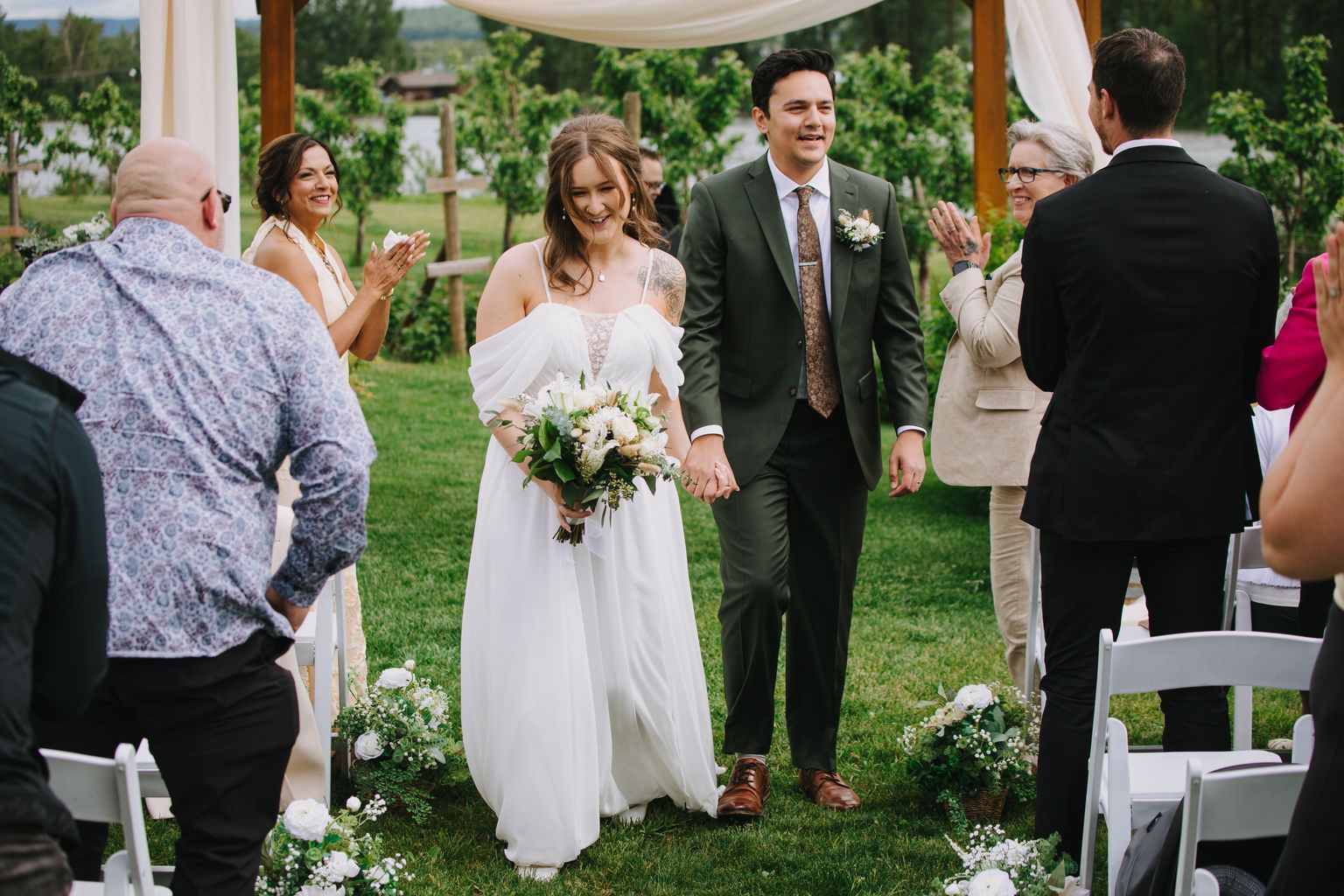 Bride in white off-shoulder gown and groom in olive suit hold hands walking across lawn with white bouquet and greenery backdrop