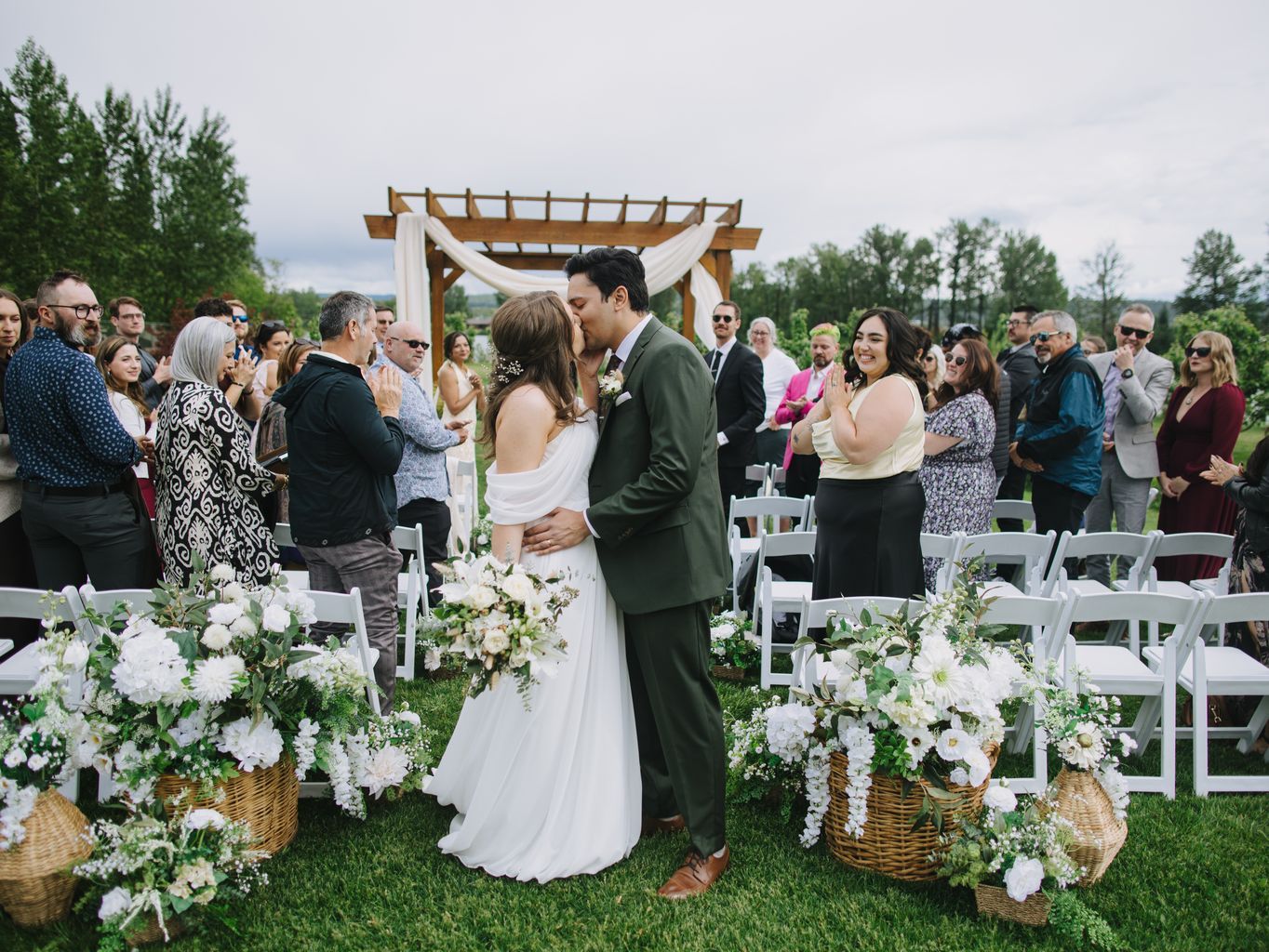 Bride and groom kiss at outdoor garden ceremony with wooden pergola, white flowers, and guests gathered beneath soft daylight