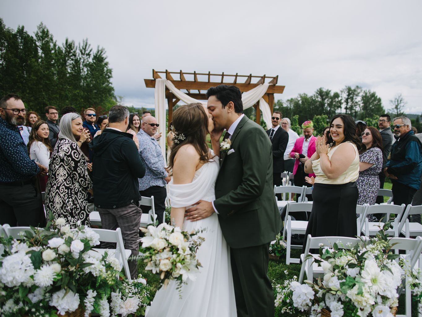 Bride and groom kiss under wooden pergola with white draping at outdoor ceremony surrounded by guests and white floral arrangements