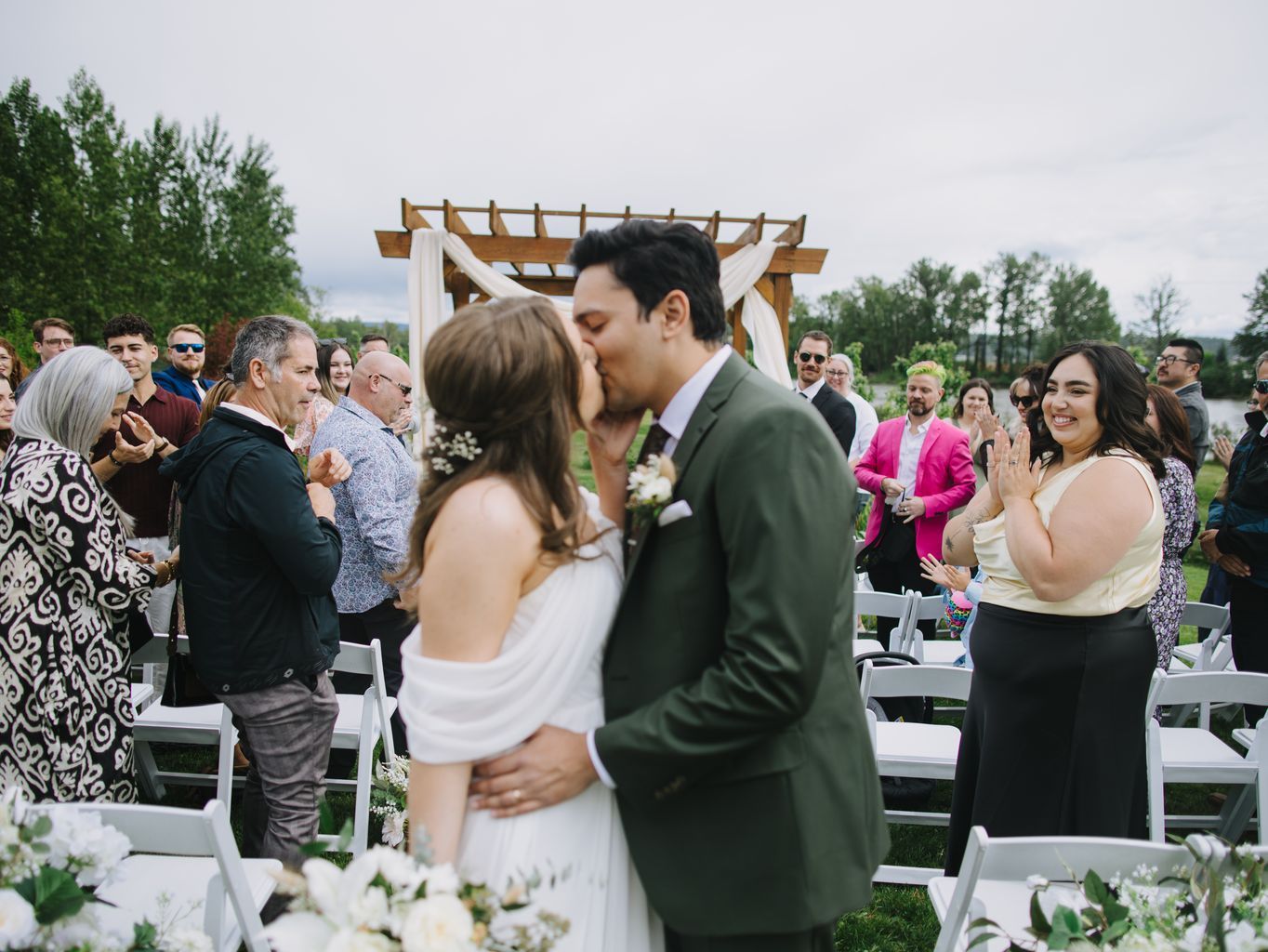Bride and groom kiss under wooden pergola with white draping during outdoor ceremony