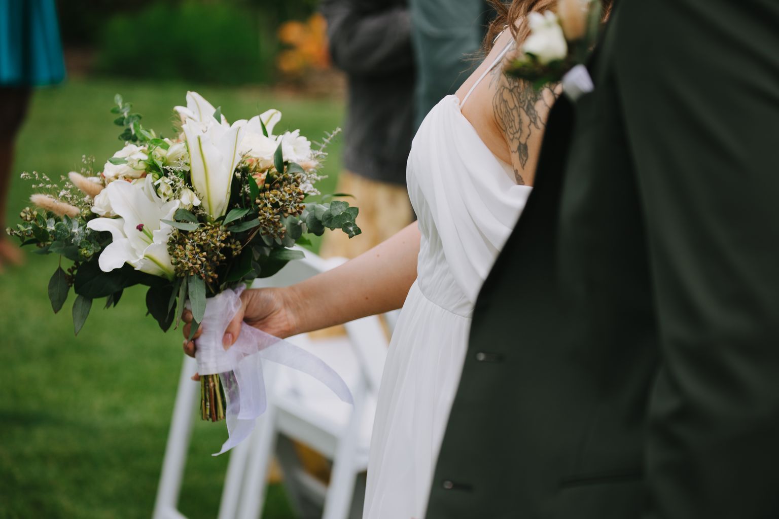 Bride in white dress holding white and green bouquet with tattooed shoulder visible, groom blurred in background