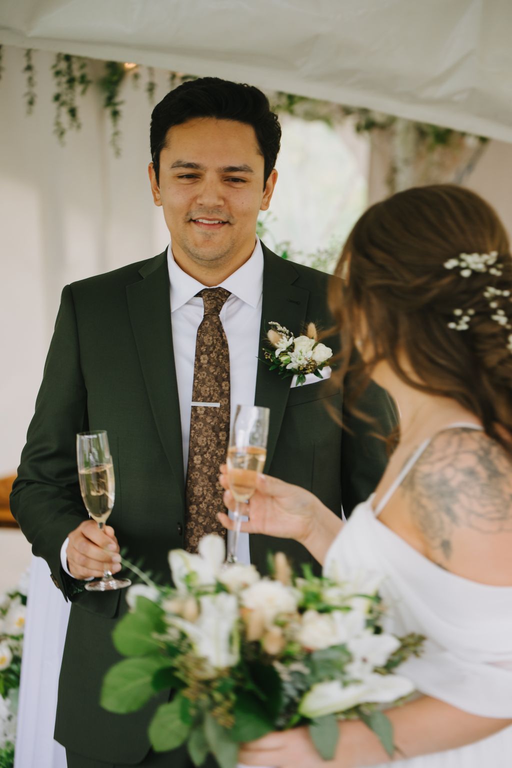Groom in dark suit and patterned tie with boutonniere smiles during wedding ceremony with bride holding champagne glass