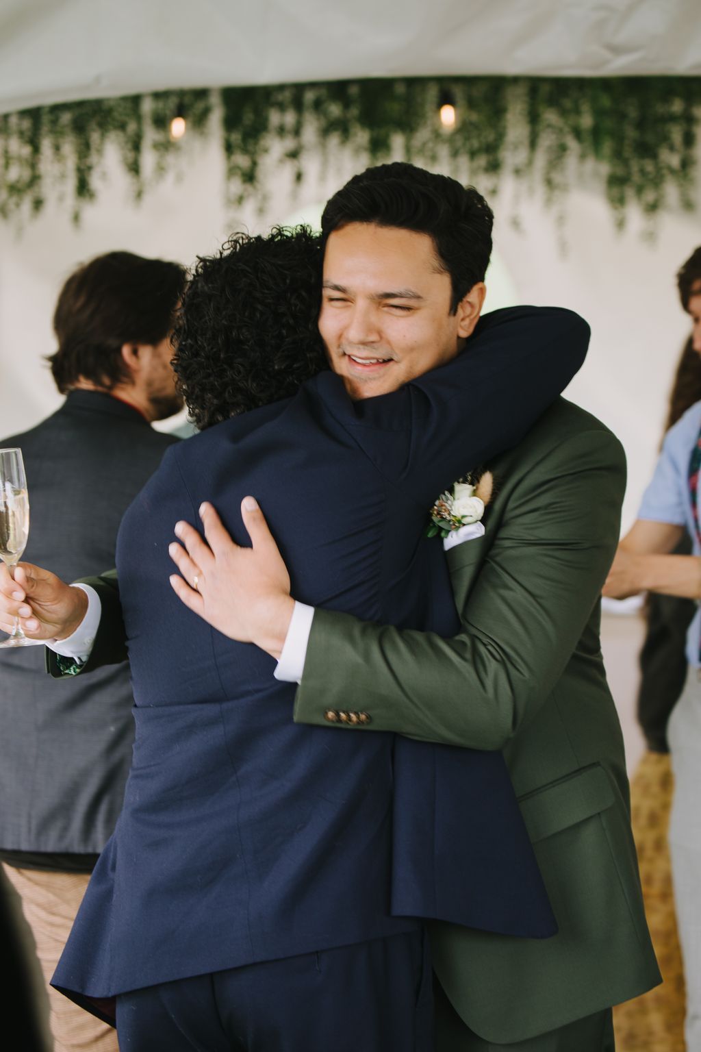 Groom in navy suit embraces bride in sage green dress, both smiling, with cascading greenery and soft lighting in background