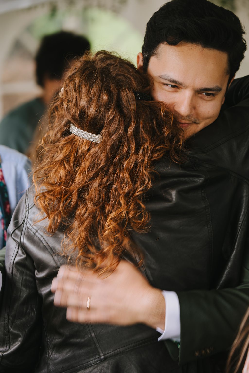 Groom in black suit embraces bride with long copper-brown curled hair and pearl headpiece, soft natural light