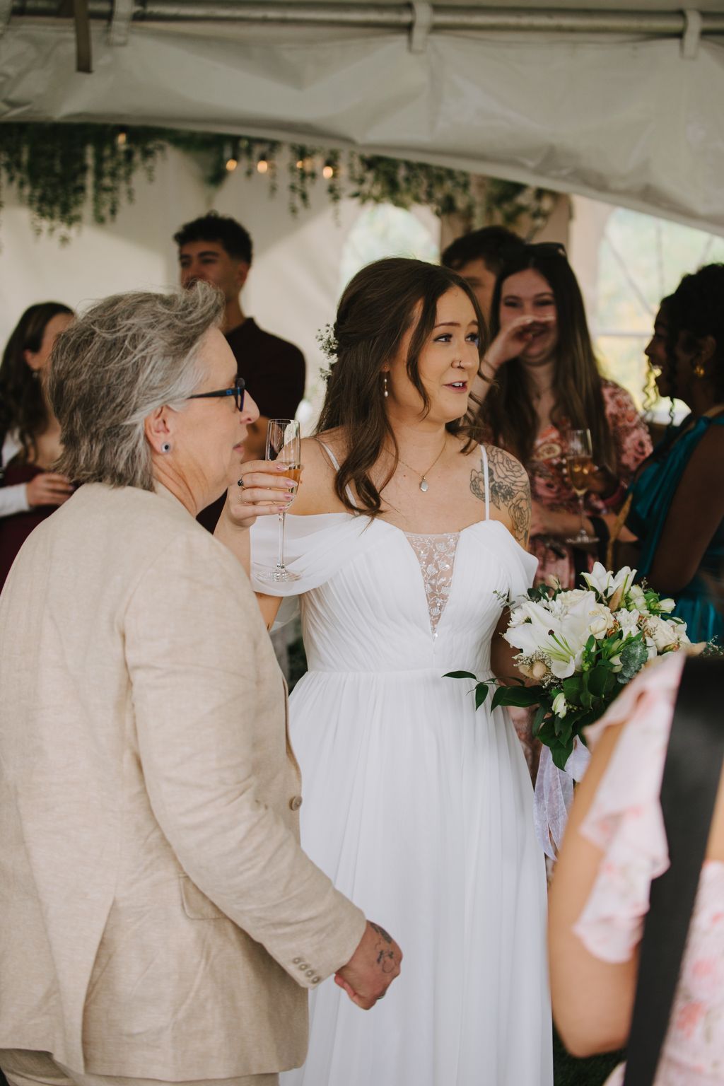 Bride in white off-shoulder dress holding champagne flute under tent with guests during reception