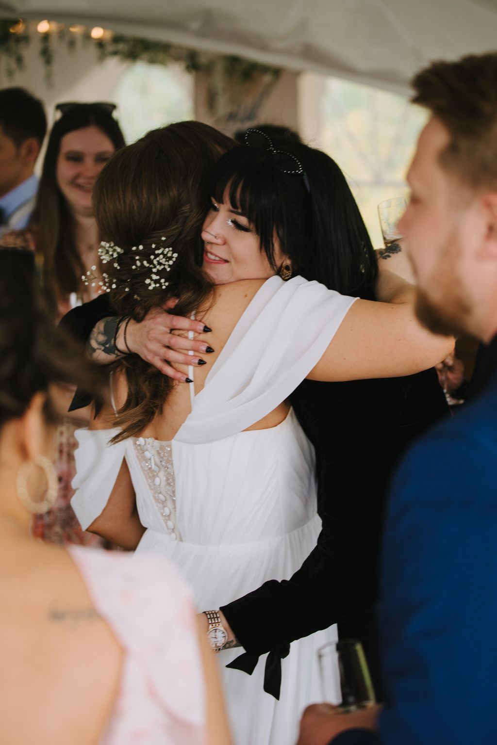 Two women embrace warmly indoors, one in a white lace dress and the other in black, sharing a joyful moment with soft natural lighting