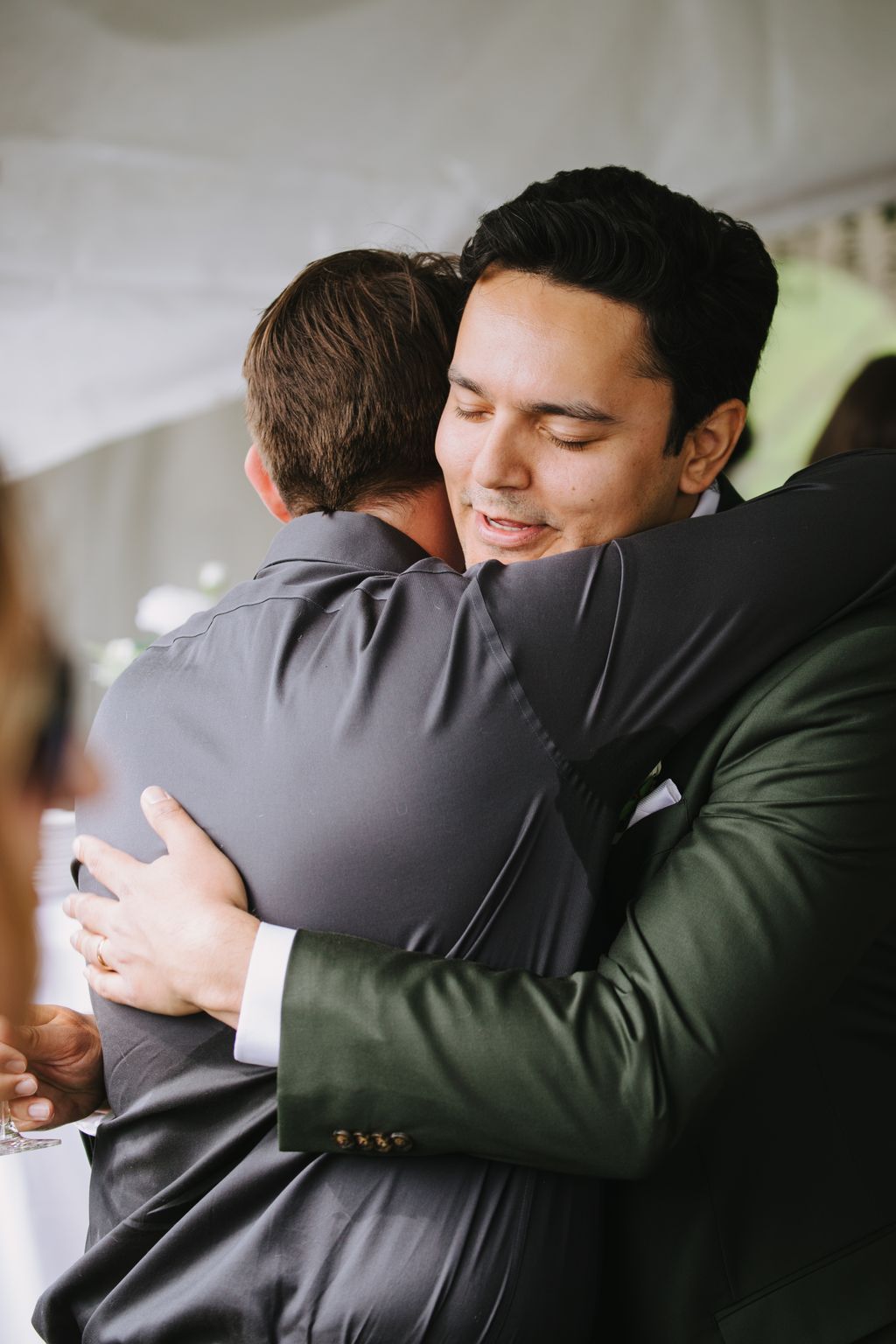 Groom in dark suit embracing best man from behind, both smiling with eyes closed in warm moment against neutral background