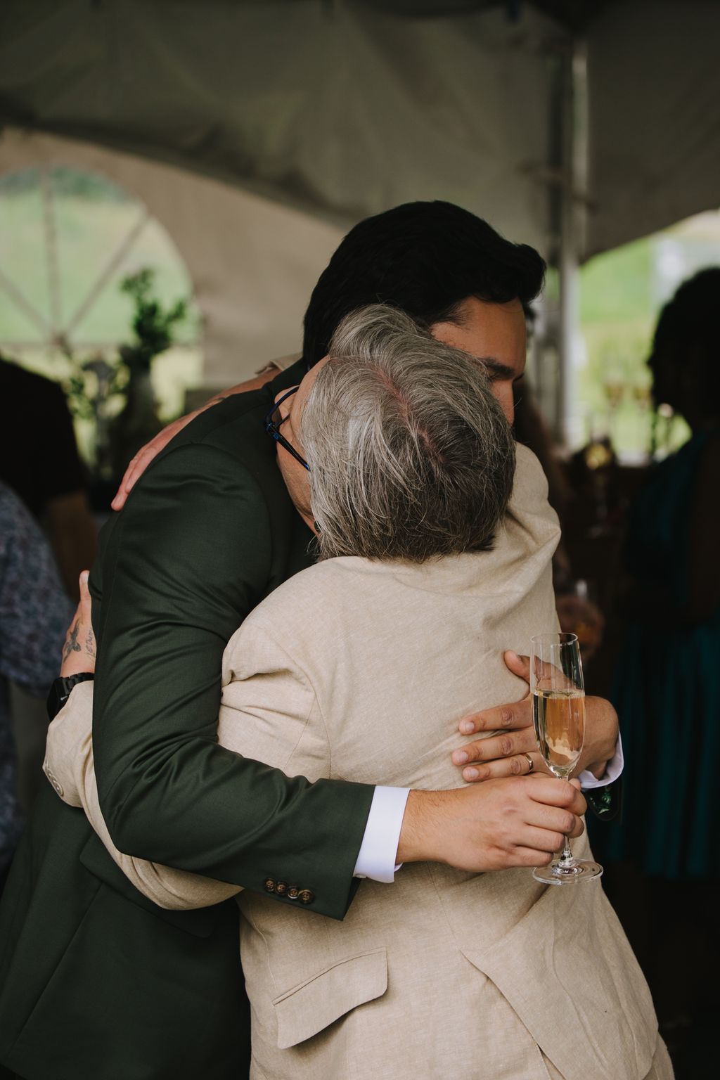 Groom in dark suit embracing elderly woman with gray hair in beige jacket, tender moment captured in soft natural light