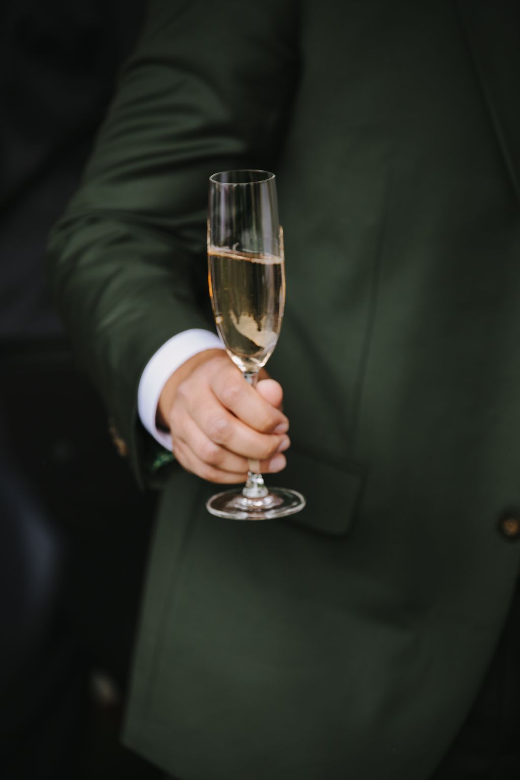 Groom in dark suit holding champagne flute against blurred green background, white dress shirt cuff visible