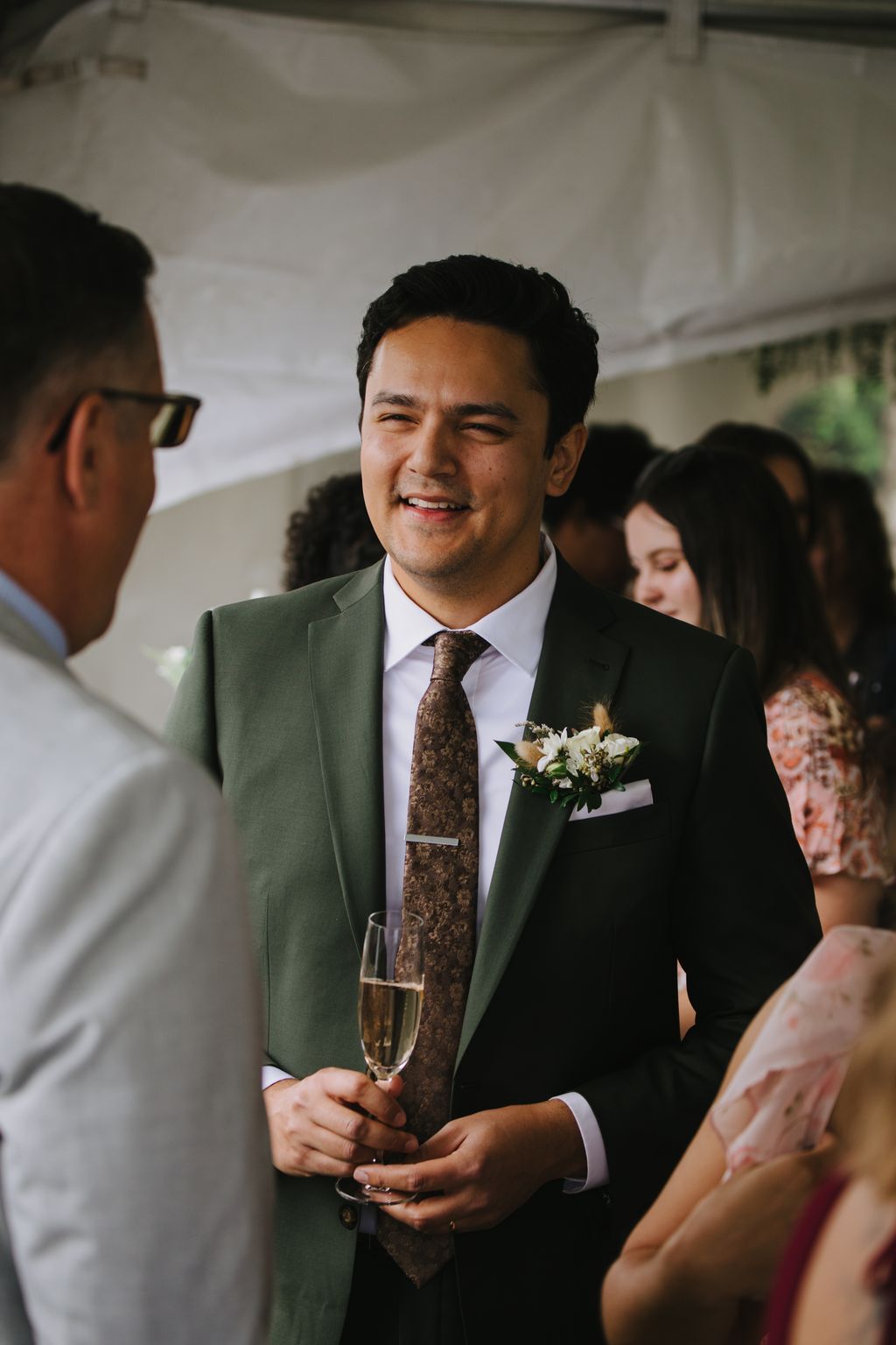 Groom in sage green suit and brown patterned tie smiles while holding champagne glass at wedding reception
