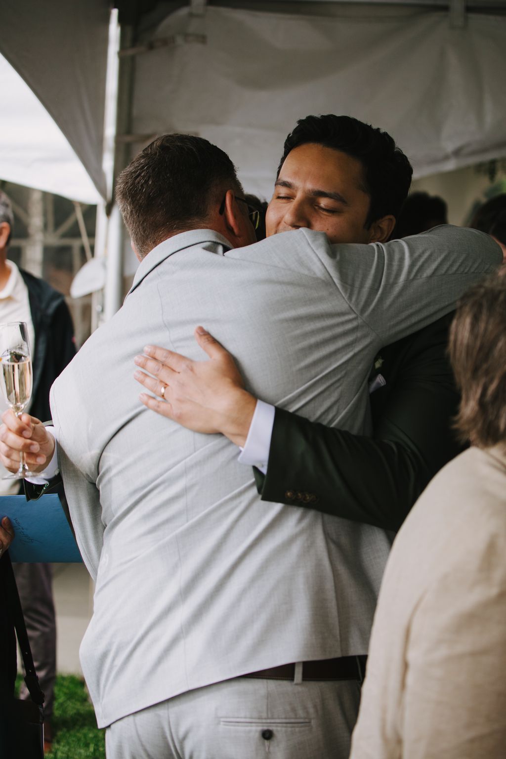 Two men embrace warmly in light-filled indoor space, one wearing a light gray suit and the other a white dress shirt, sharing an intimate moment of connection