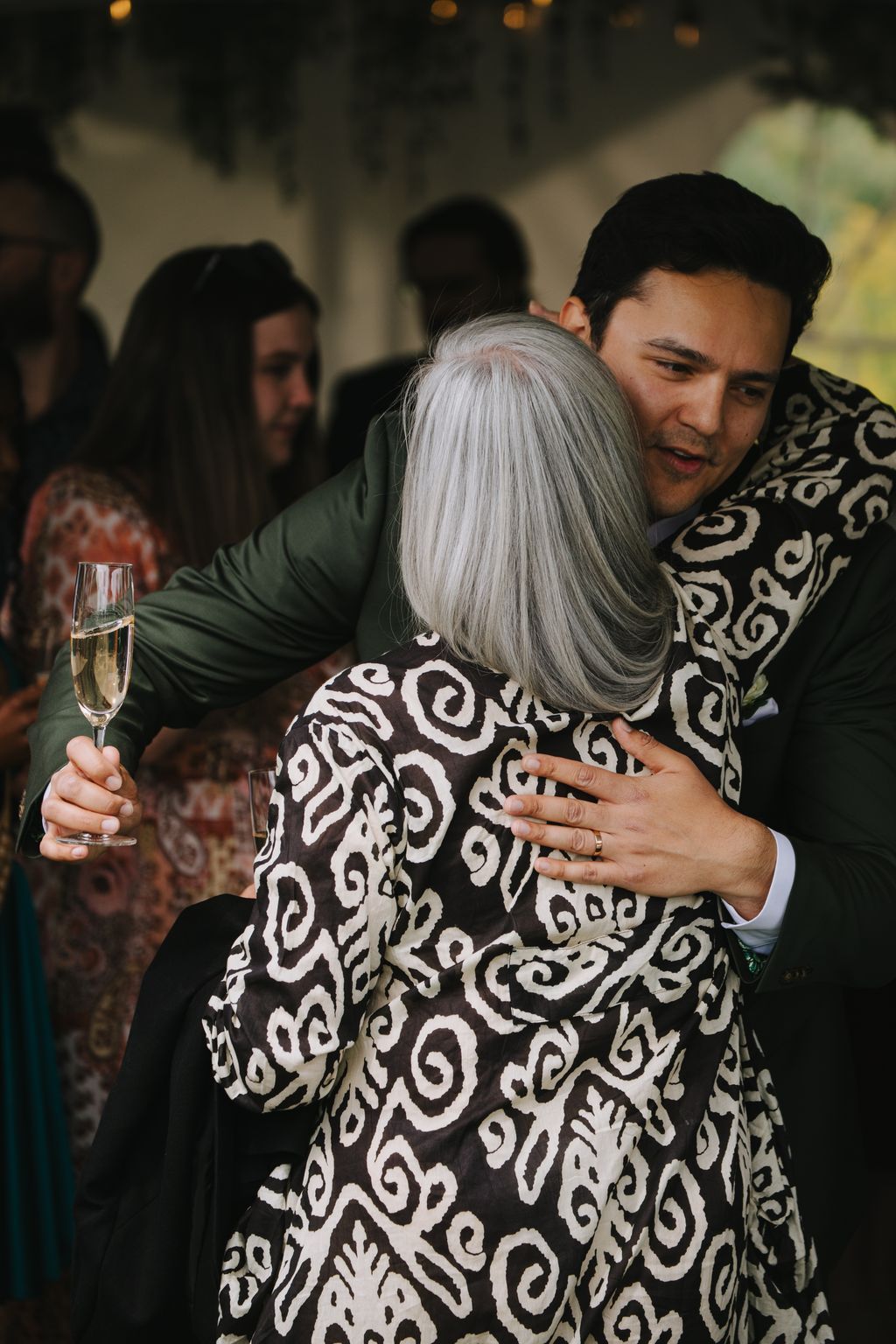 Groom embraces bride from behind in patterned dress, warm indoor lighting, intimate moment with guests blurred in background