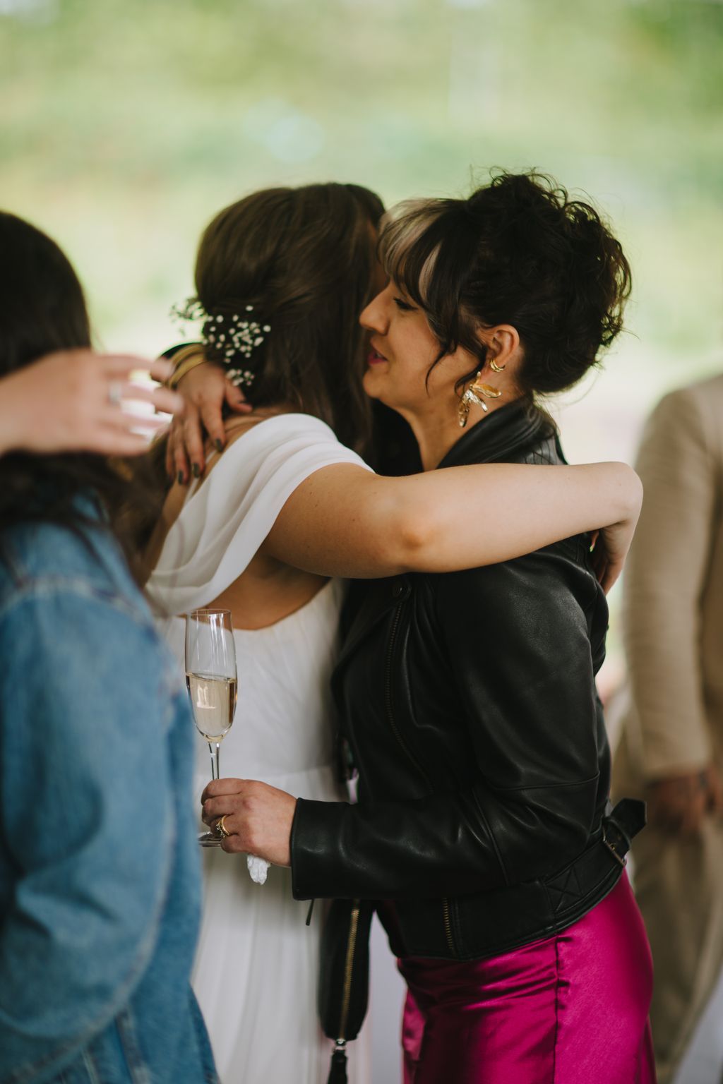 Two women embrace while holding champagne, one wearing white and one in black leather, with baby''s breath in her hair against a soft blurred background
