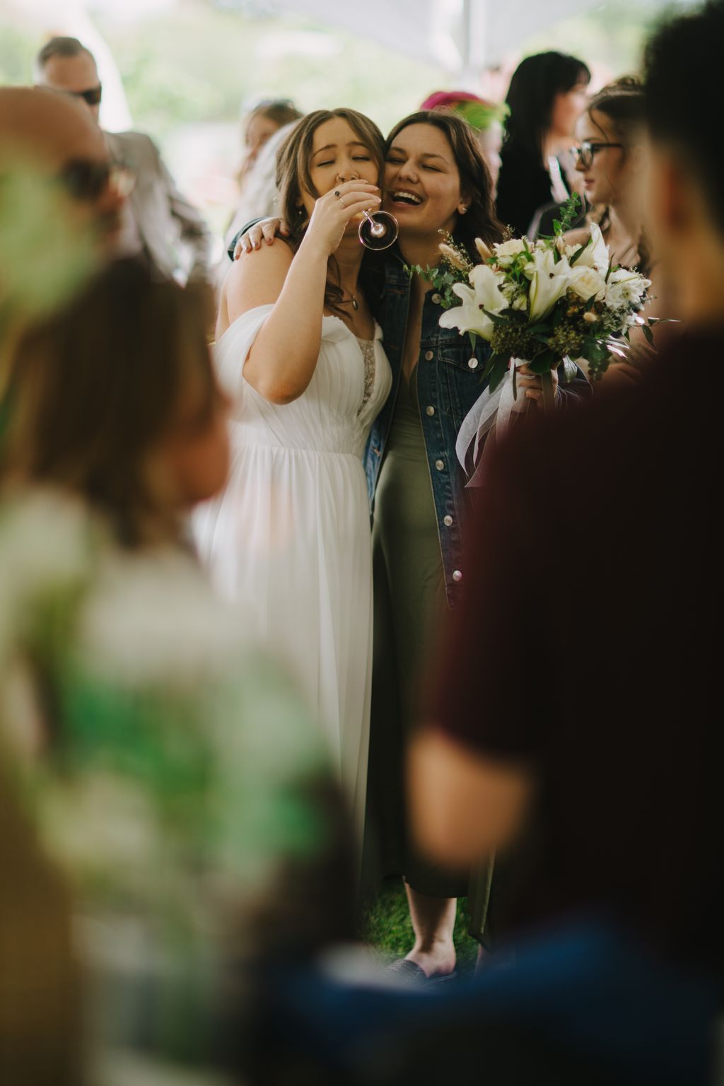 Two women laugh joyfully together outdoors, one in a white dress holding white flowers, surrounded by wedding guests in soft natural light