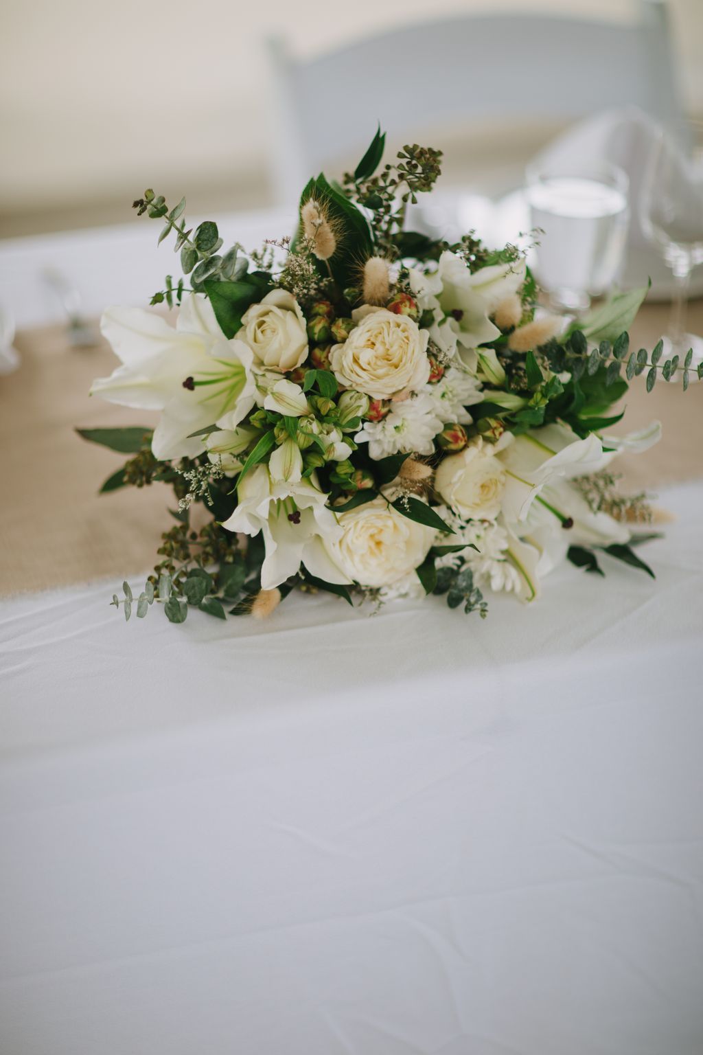 Romantic bridal bouquet with cream roses, white flowers, green foliage, and textured greenery on soft white fabric