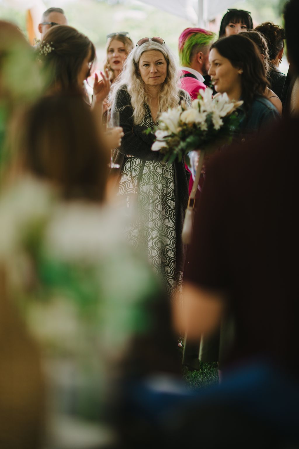 Woman with long white hair and sunglasses surrounded by guests at an outdoor wedding ceremony, holding white flowers
