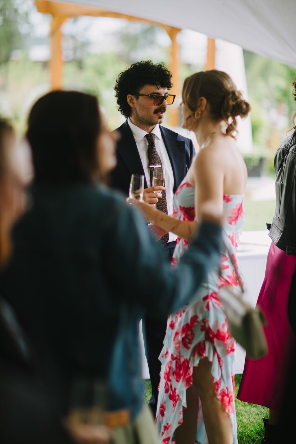 Groom in navy suit and orange-tinted glasses holds champagne while conversing with guests under a wooden pergola at an outdoor reception