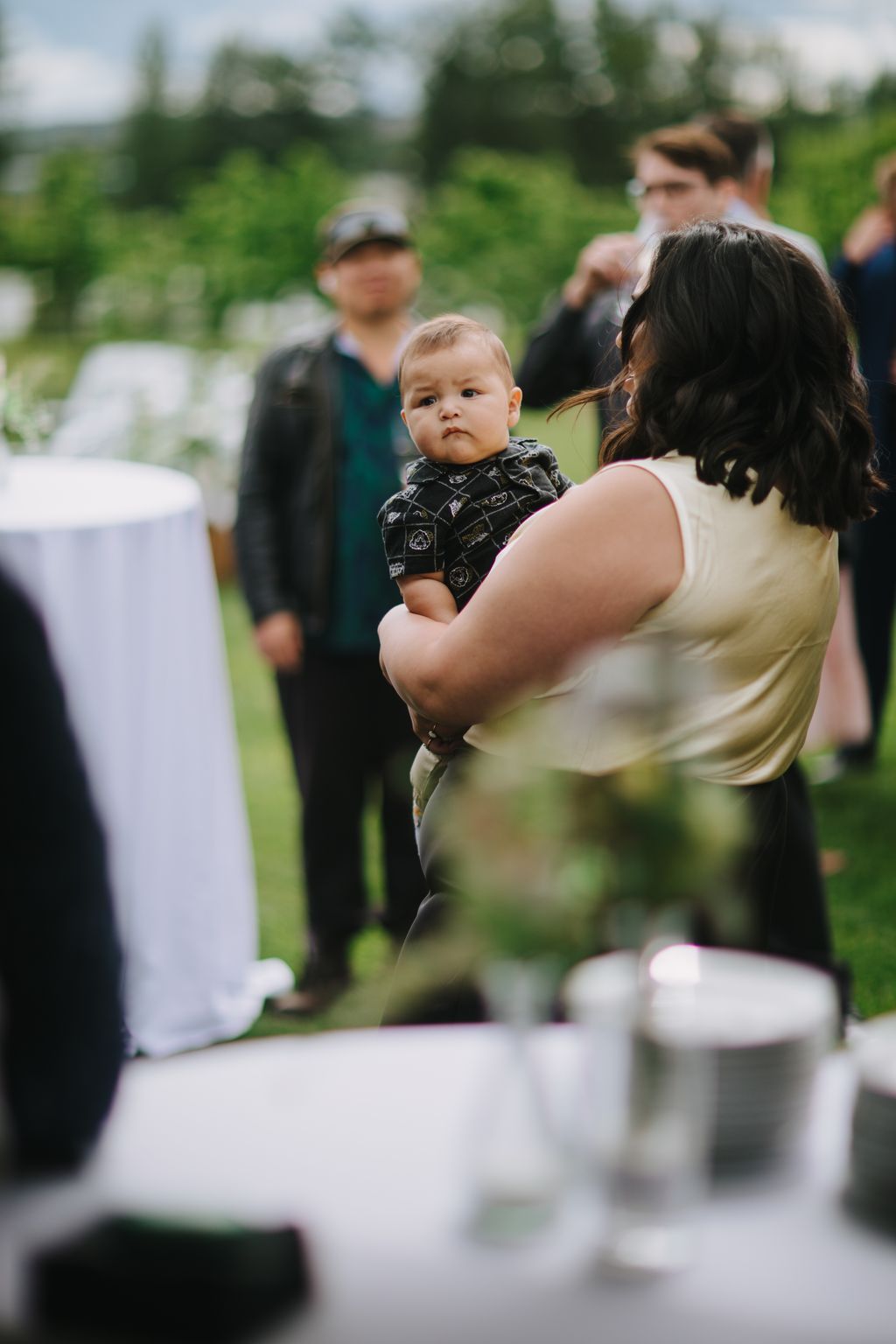 Toddler in patterned shirt held by woman at outdoor garden wedding ceremony with guests blurred in background