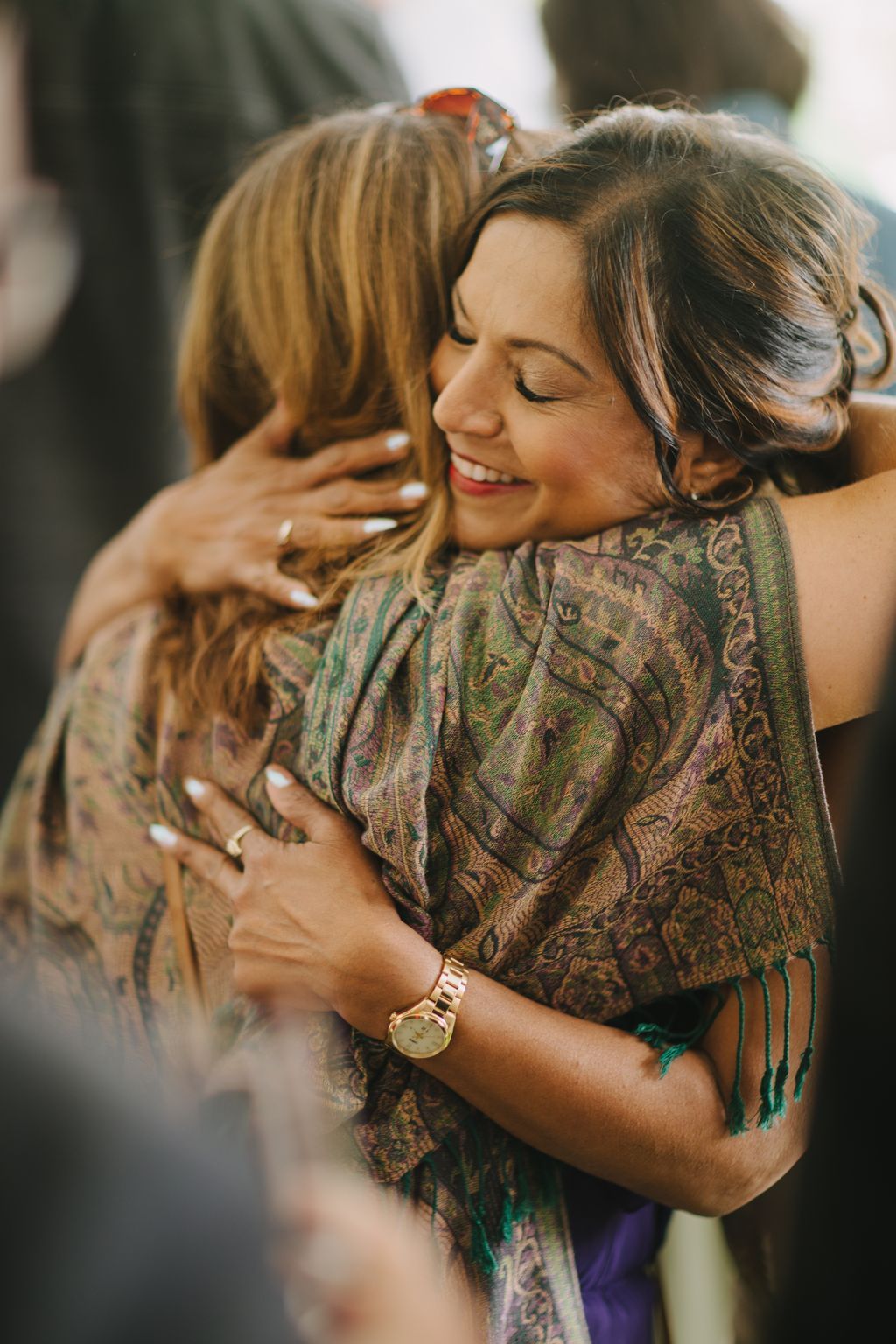 Two women embrace joyfully outdoors, one wearing a patterned green and gold jacket, with natural daylight and soft focus foliage in the background