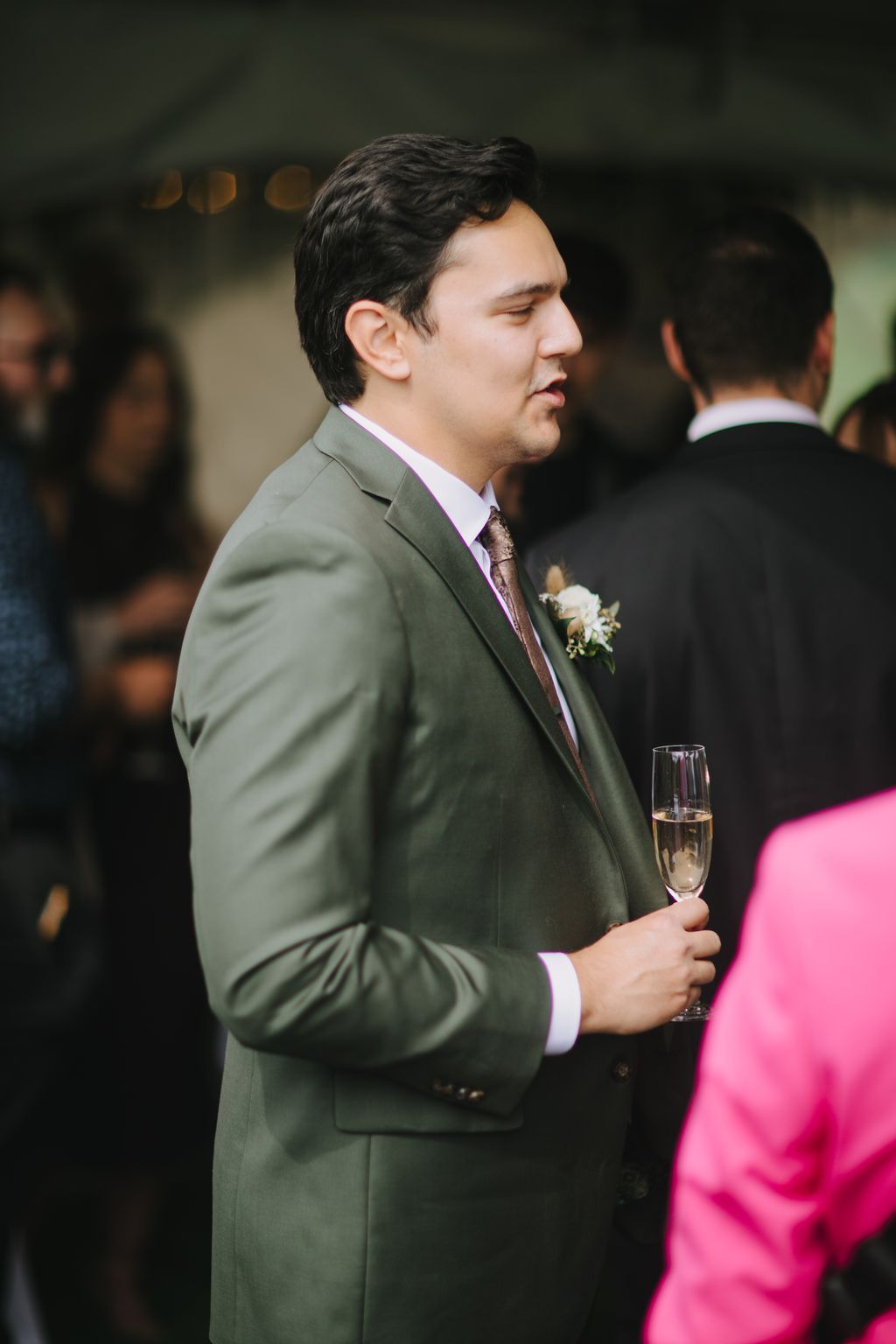 Groom in sage green suit with lavender shirt and floral boutonniere gazes thoughtfully to the side during wedding ceremony