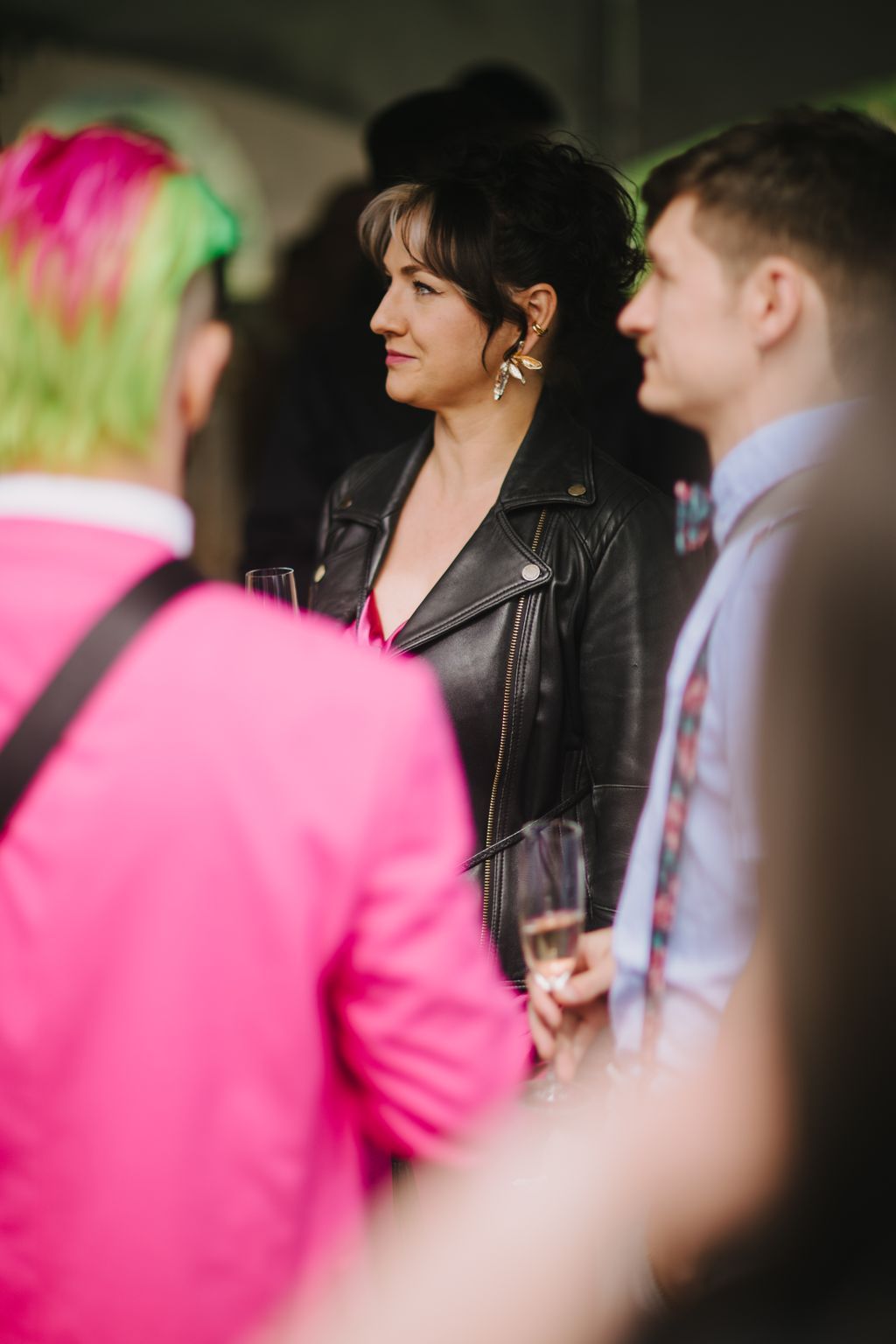 Woman in black leather jacket and statement earrings at an evening event, holding a glass with pink-clad guests nearby