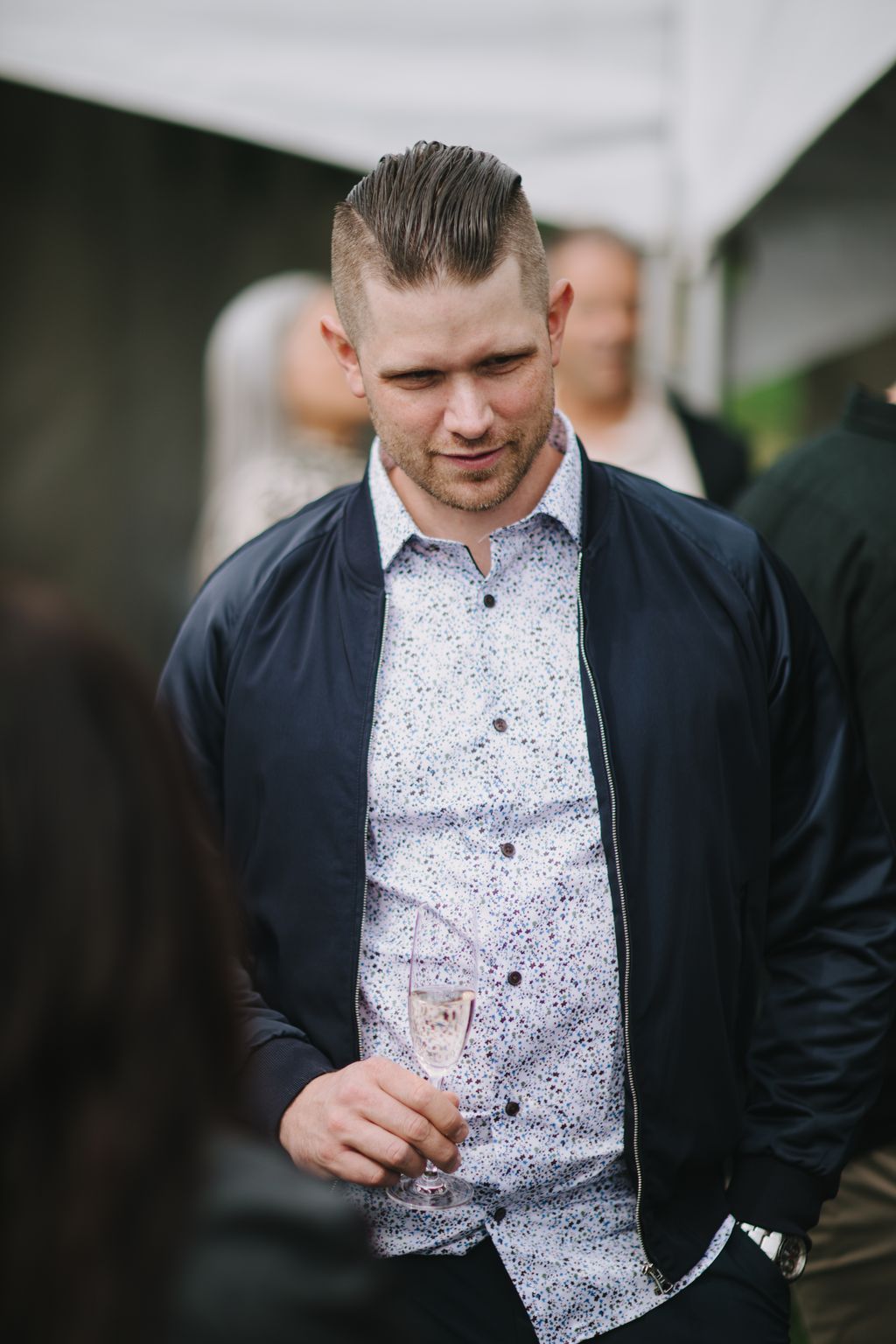 Groom in navy jacket and patterned shirt holds champagne flute, smiling downward in outdoor daylight