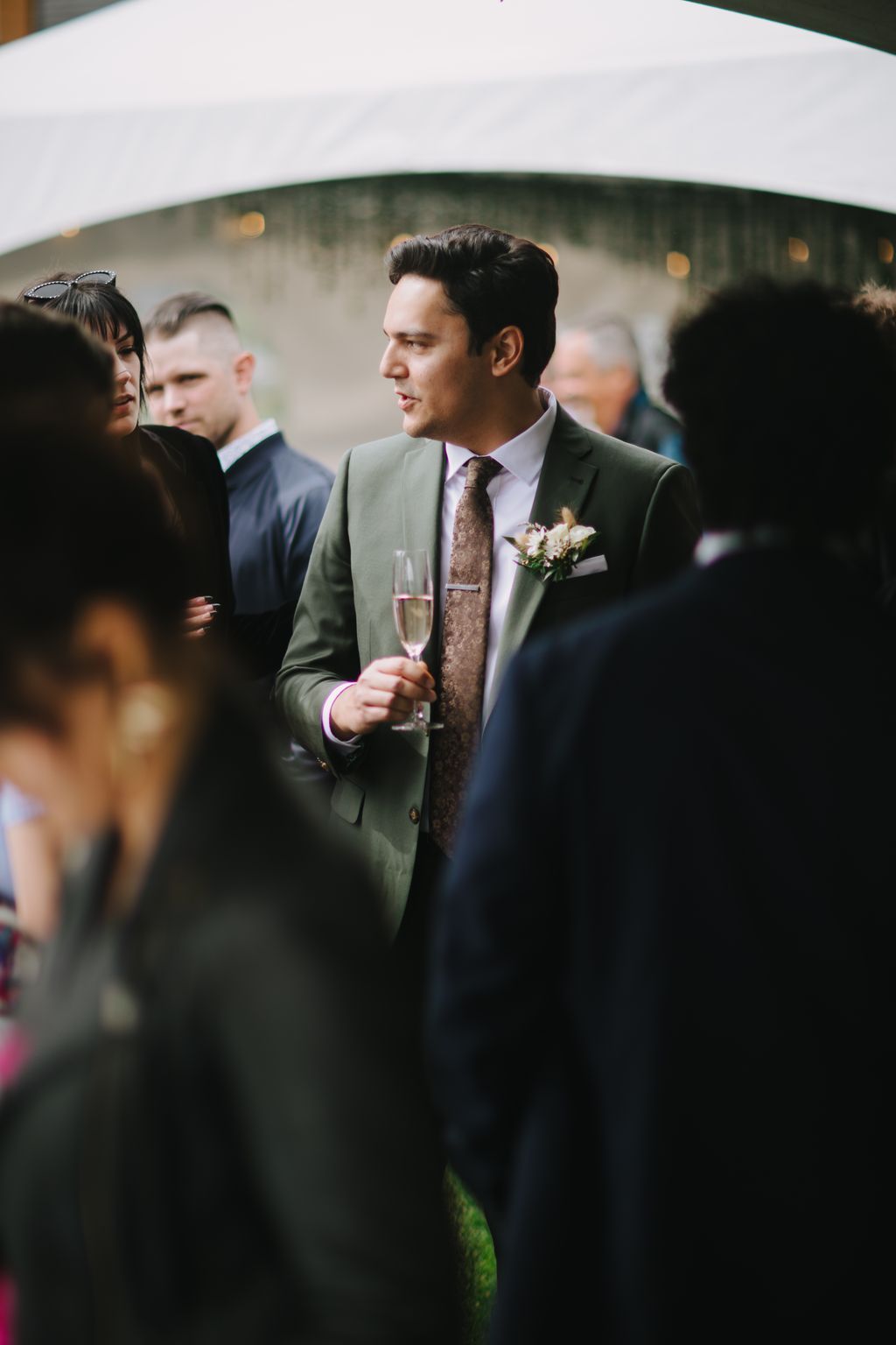 Groom in sage green suit holding champagne flute at outdoor wedding reception, smiling to the side