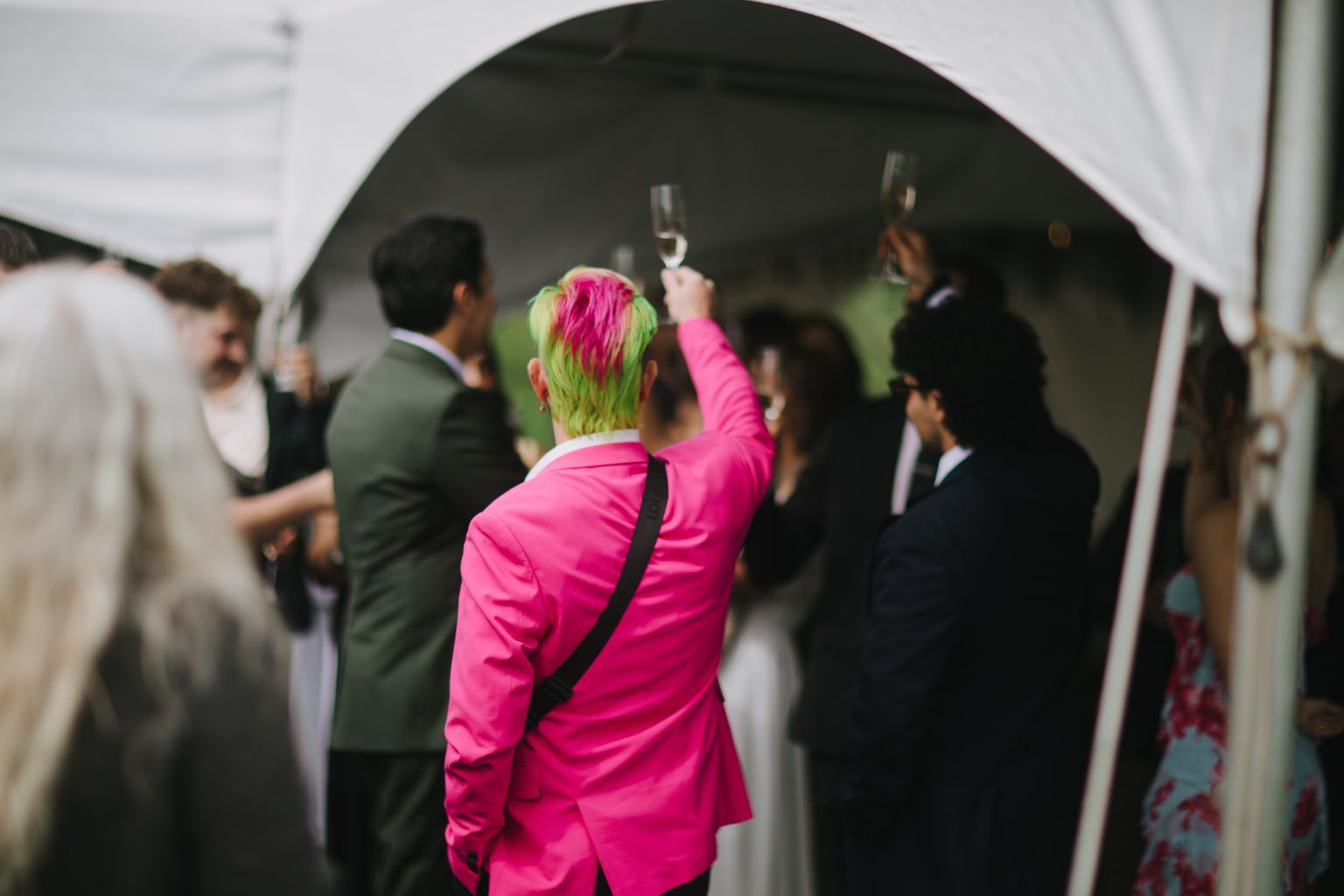 Person in vibrant pink blazer with green and magenta hair raises a glass at an indoor celebration surrounded by guests