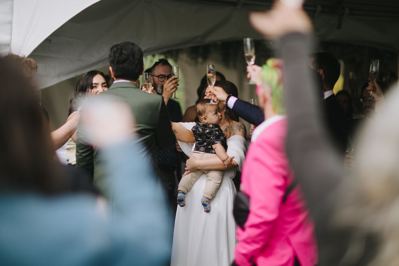 Bride in white dress holds toddler while guests toast with champagne under tent at wedding celebration