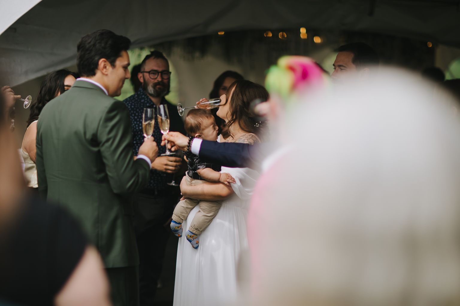 Bride holding toddler drinks champagne while groom smiles beside them at evening reception