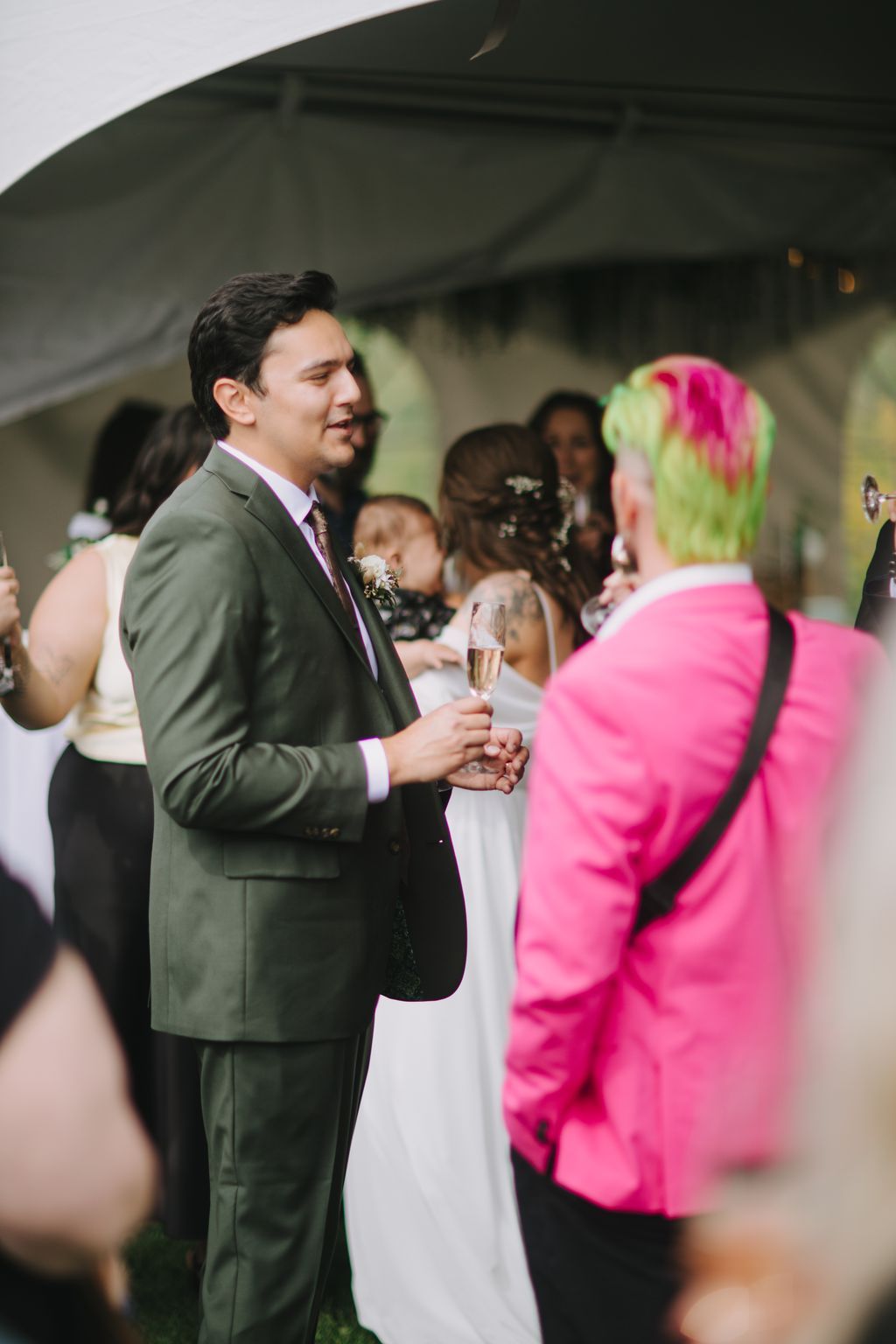 Groom in dark suit holding champagne flute smiles during outdoor reception with guests in background