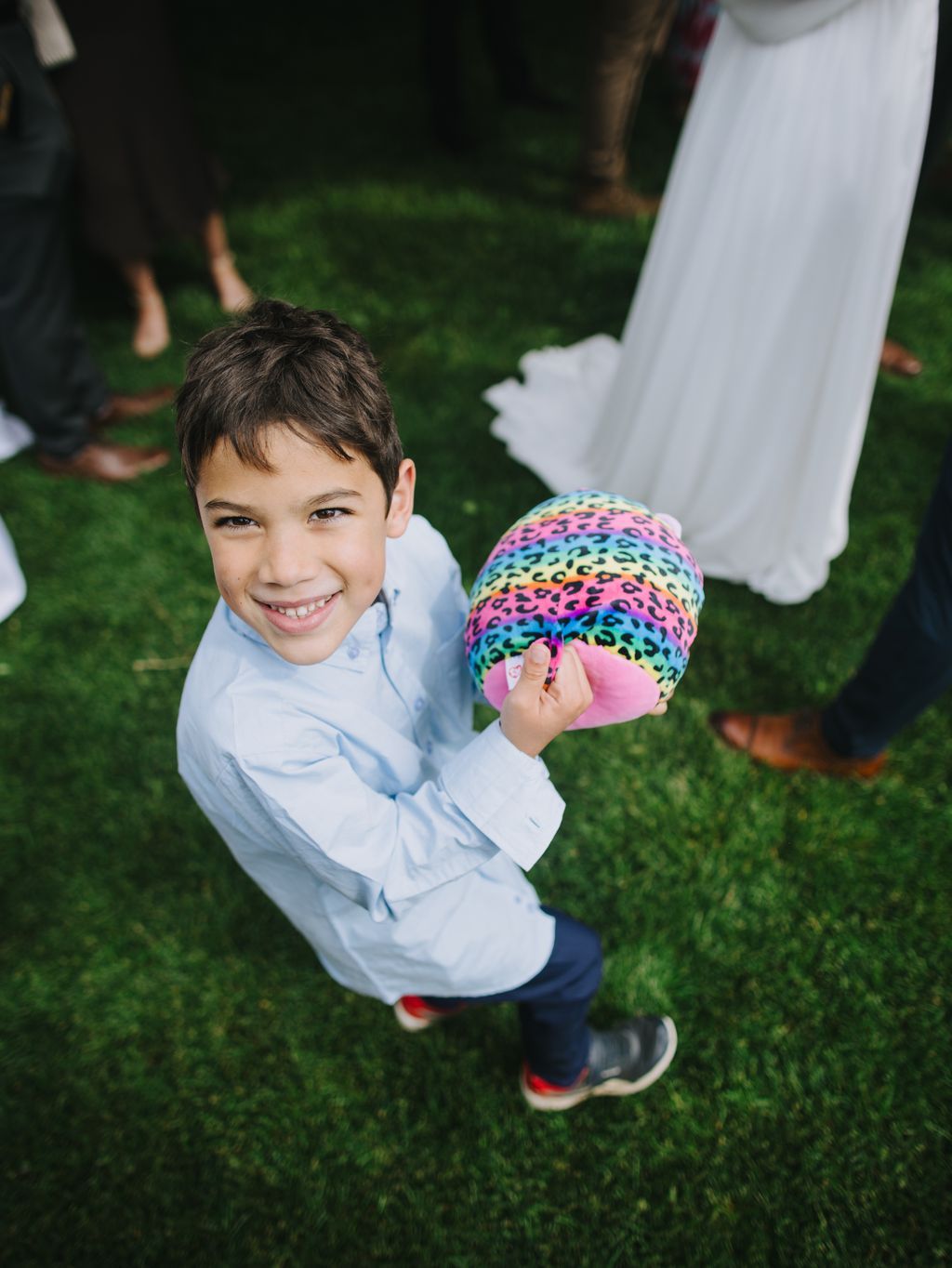 Young boy in light blue shirt smiling at camera while holding colorful rainbow leopard print ball on green lawn