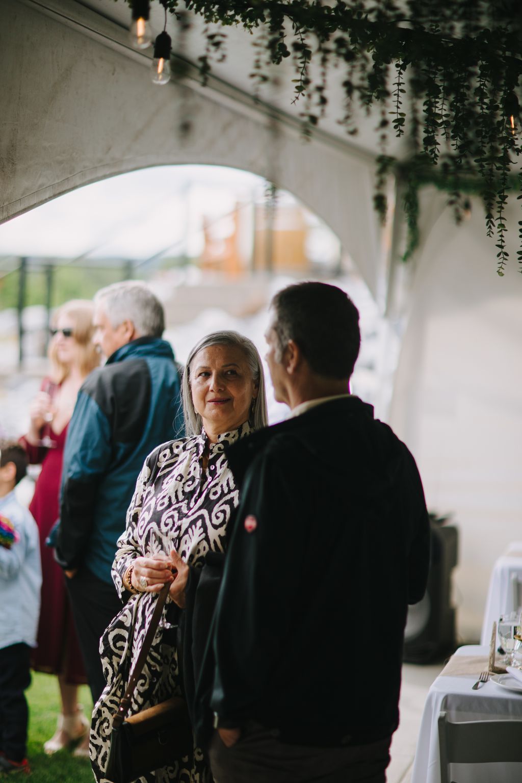 Woman in patterned white and black jacket smiles while conversing with man in dark suit beneath hanging greenery and Edison bulb