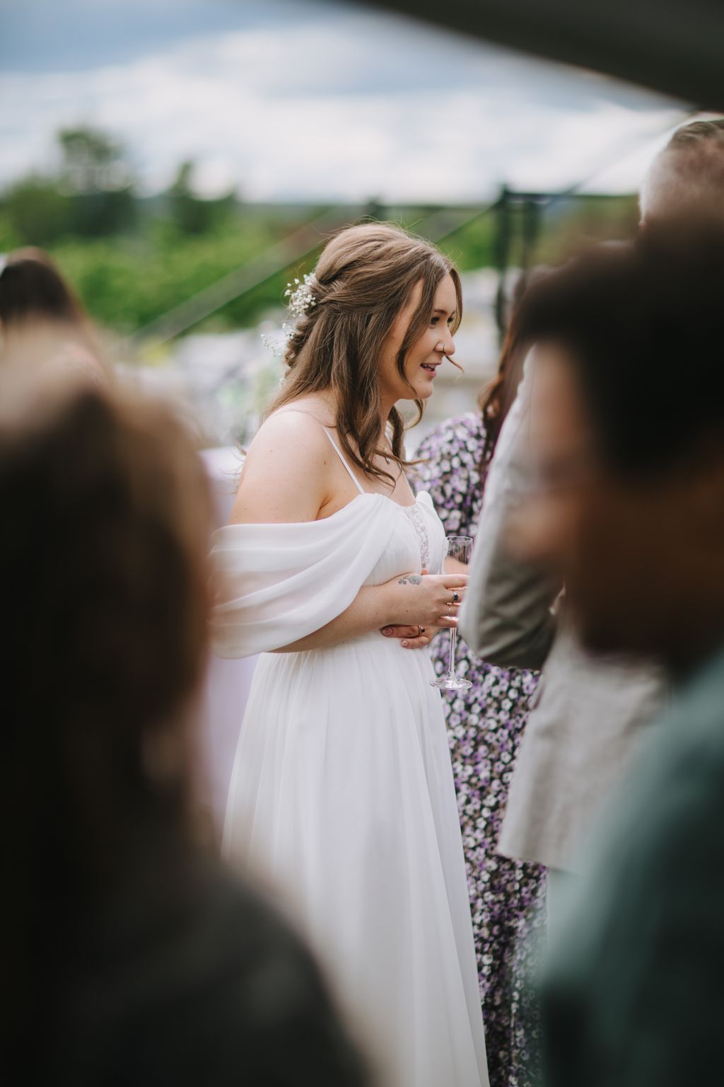 Bride in flowing white dress with baby''s breath hair piece holds champagne flute during outdoor garden ceremony with purple flowers and green fields