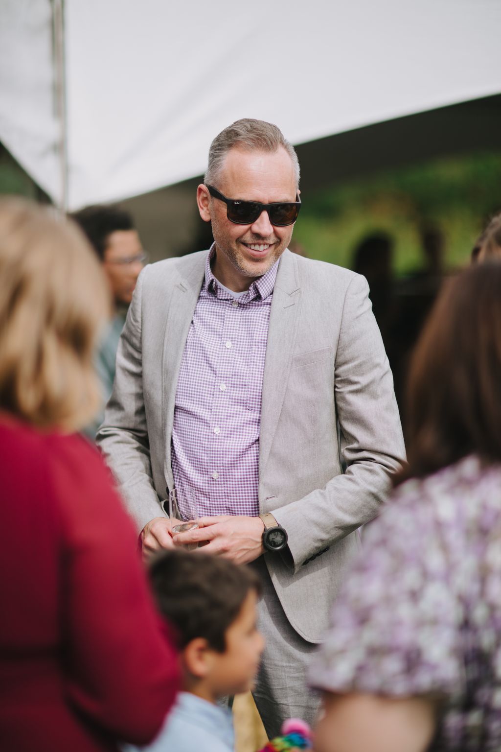 Groom in beige blazer and purple gingham shirt smiles warmly at outdoor wedding reception