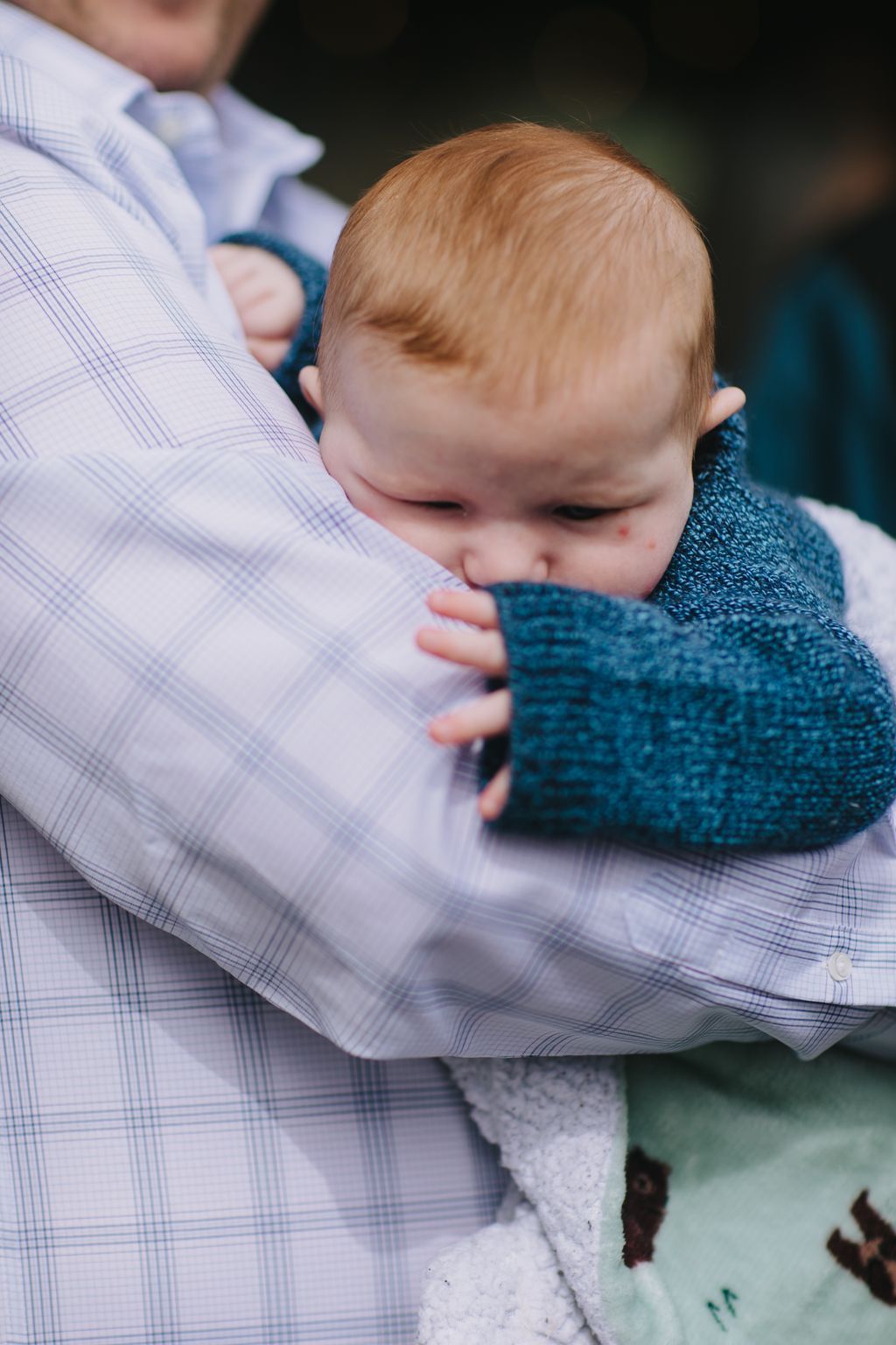 Infant with reddish-blonde hair rests on parent''s shoulder wearing blue knit sweater in soft natural light