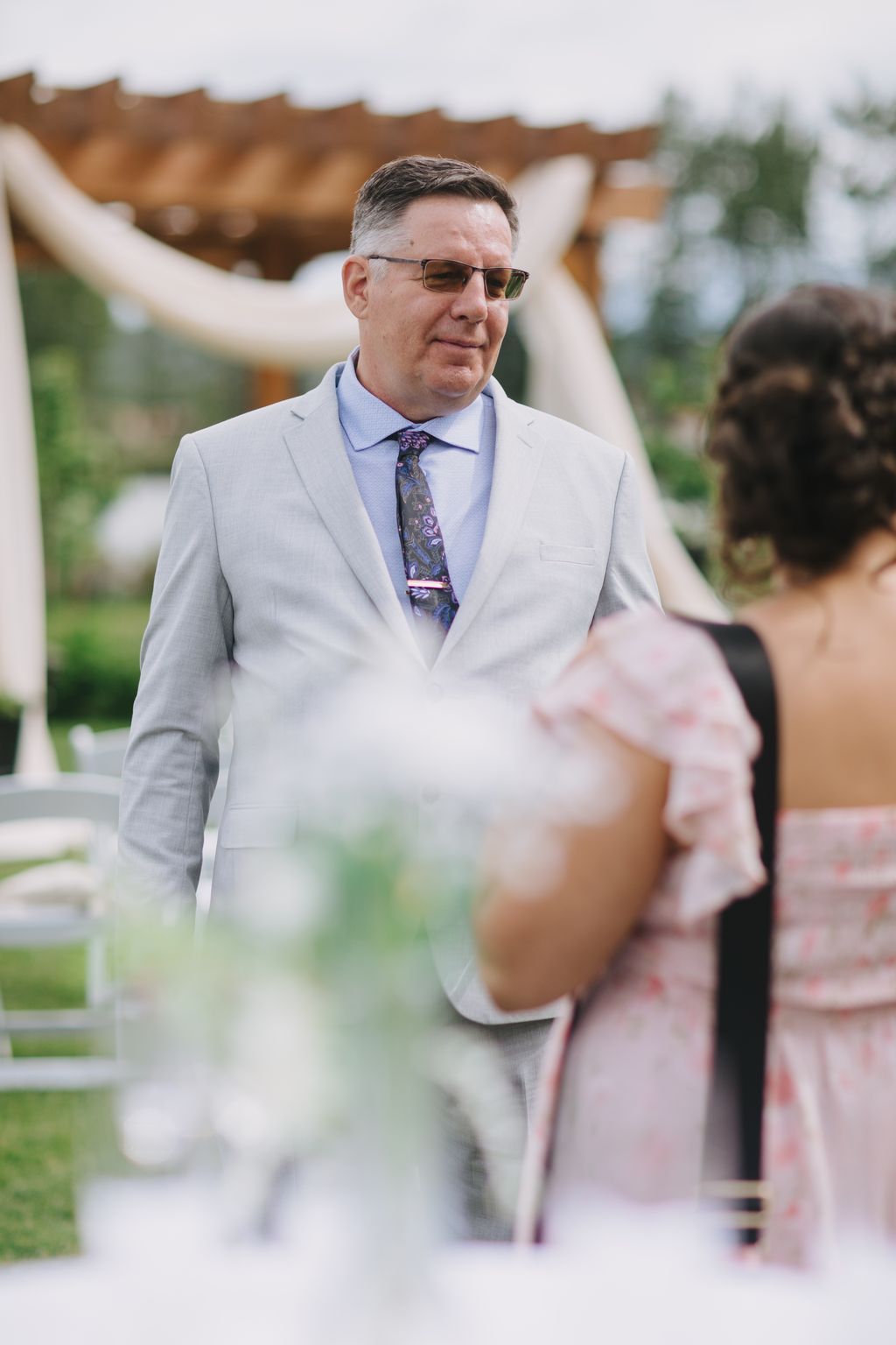Groom in light gray suit and sunglasses gazes thoughtfully during outdoor wedding ceremony