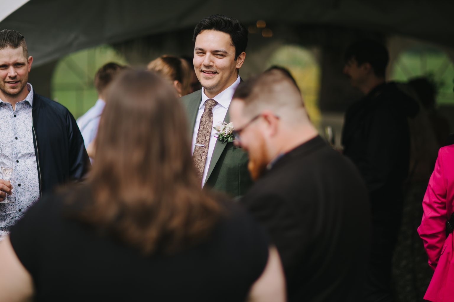 Groom in lavender shirt and patterned tie smiles warmly at wedding reception surrounded by guests