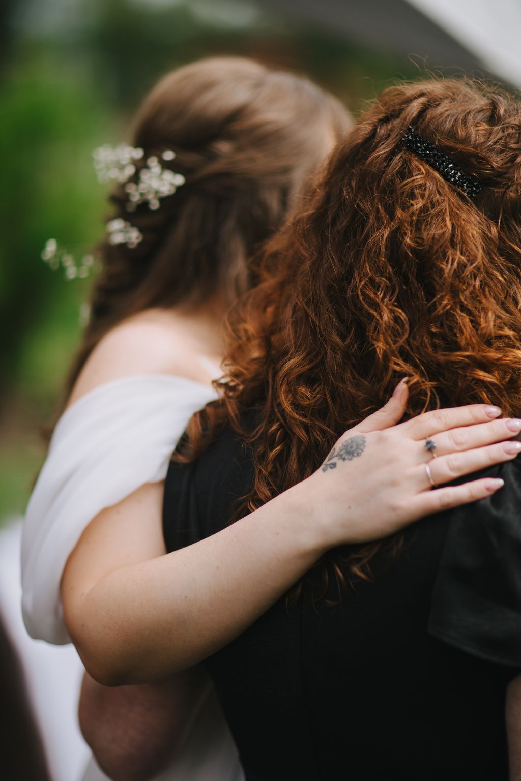 Bride with long auburn curls and small white flowers in hair embraces groom from behind in soft natural light