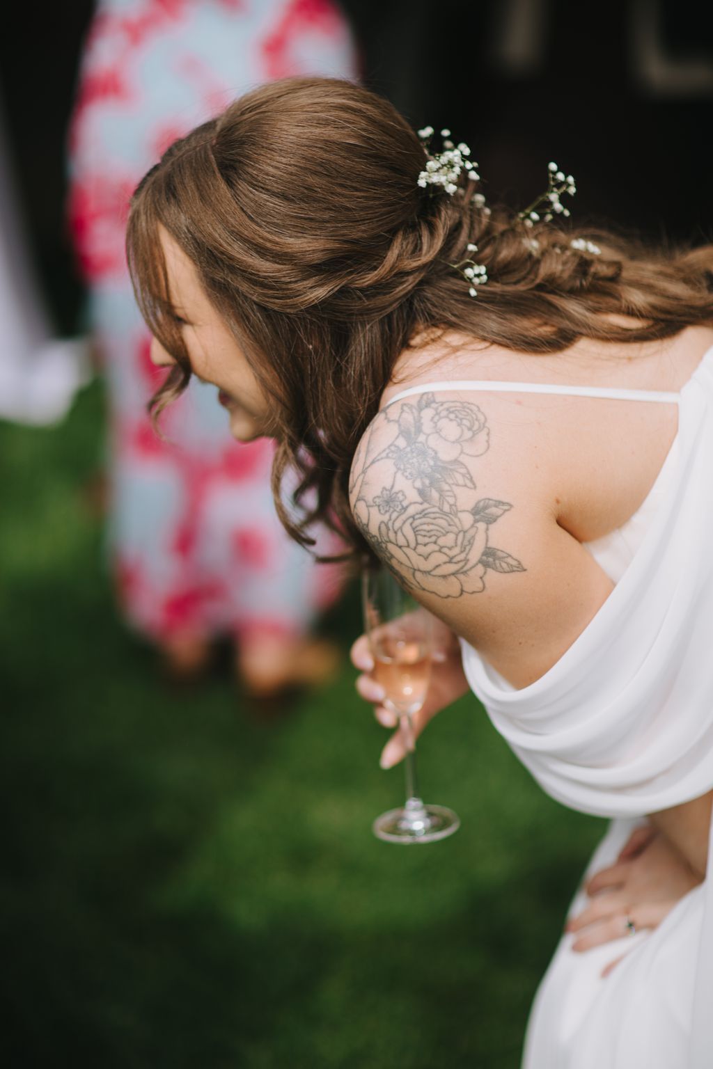 Bride with half-up brown hair adorned with baby''s breath holds champagne flute, displaying shoulder tattoo, outdoor garden setting
