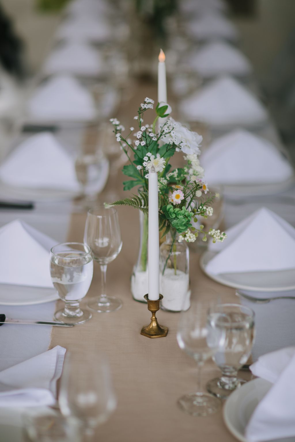 Elegant table setting with white taper candle in brass candlestick surrounded by delicate white and yellow daisy flowers and baby''s breath in a clear glass vase