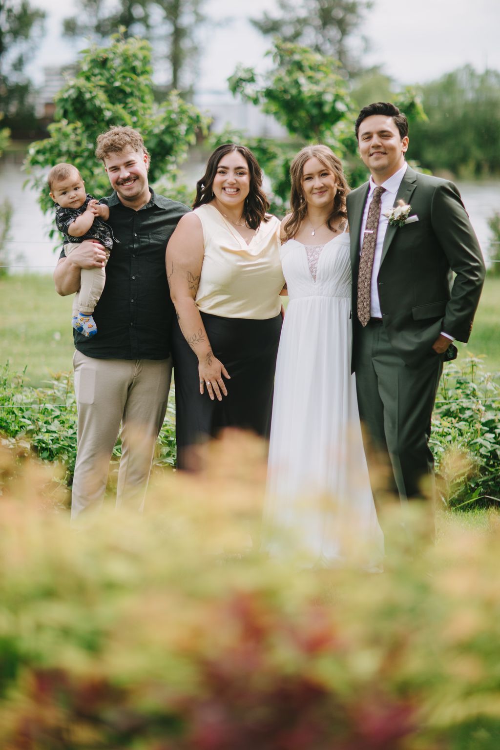 Four smiling guests in semi-formal attire stand together in a lush garden, with soft natural light filtering through green foliage behind them