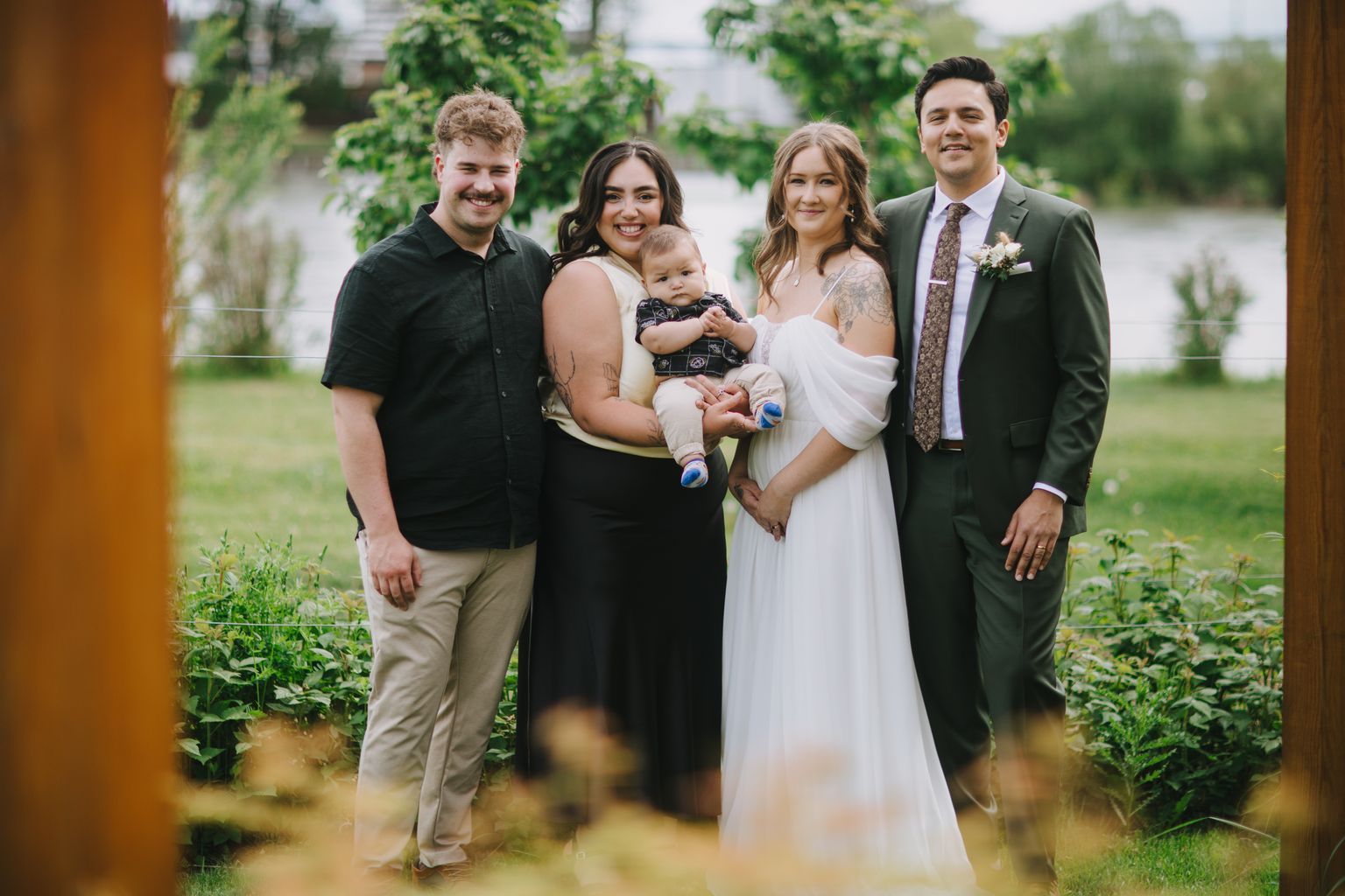 Four smiling people including a toddler pose outdoors among green foliage in soft natural light
