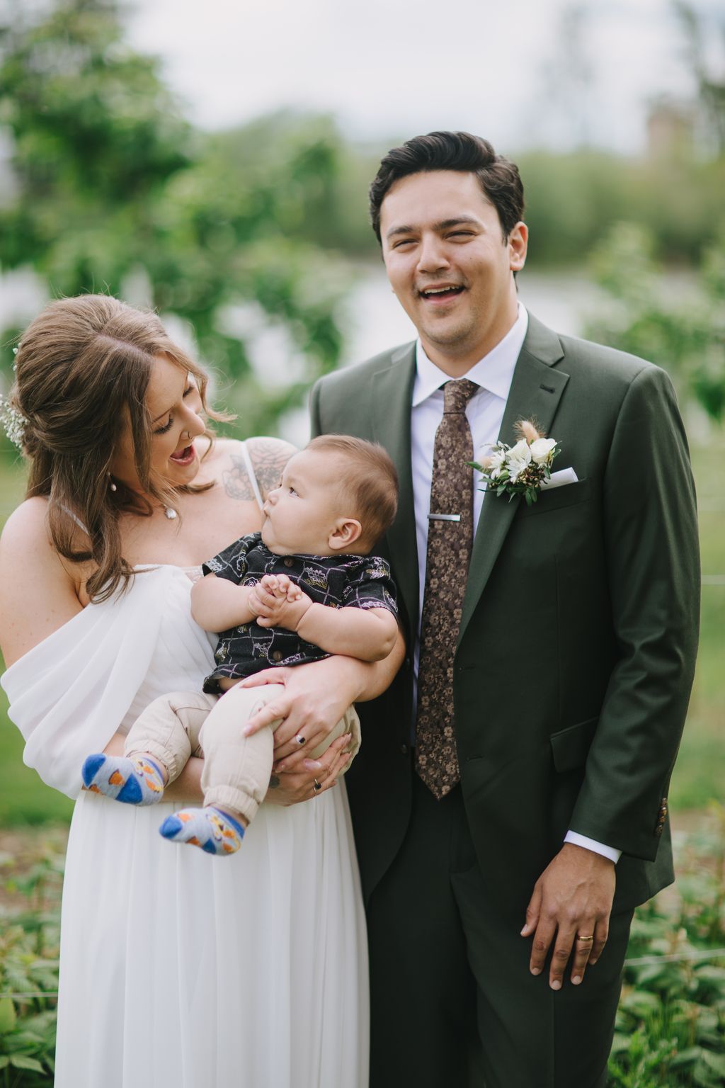 Groom in gray suit and patterned tie holds smiling infant while standing beside bride in white dress outdoors with blurred green background