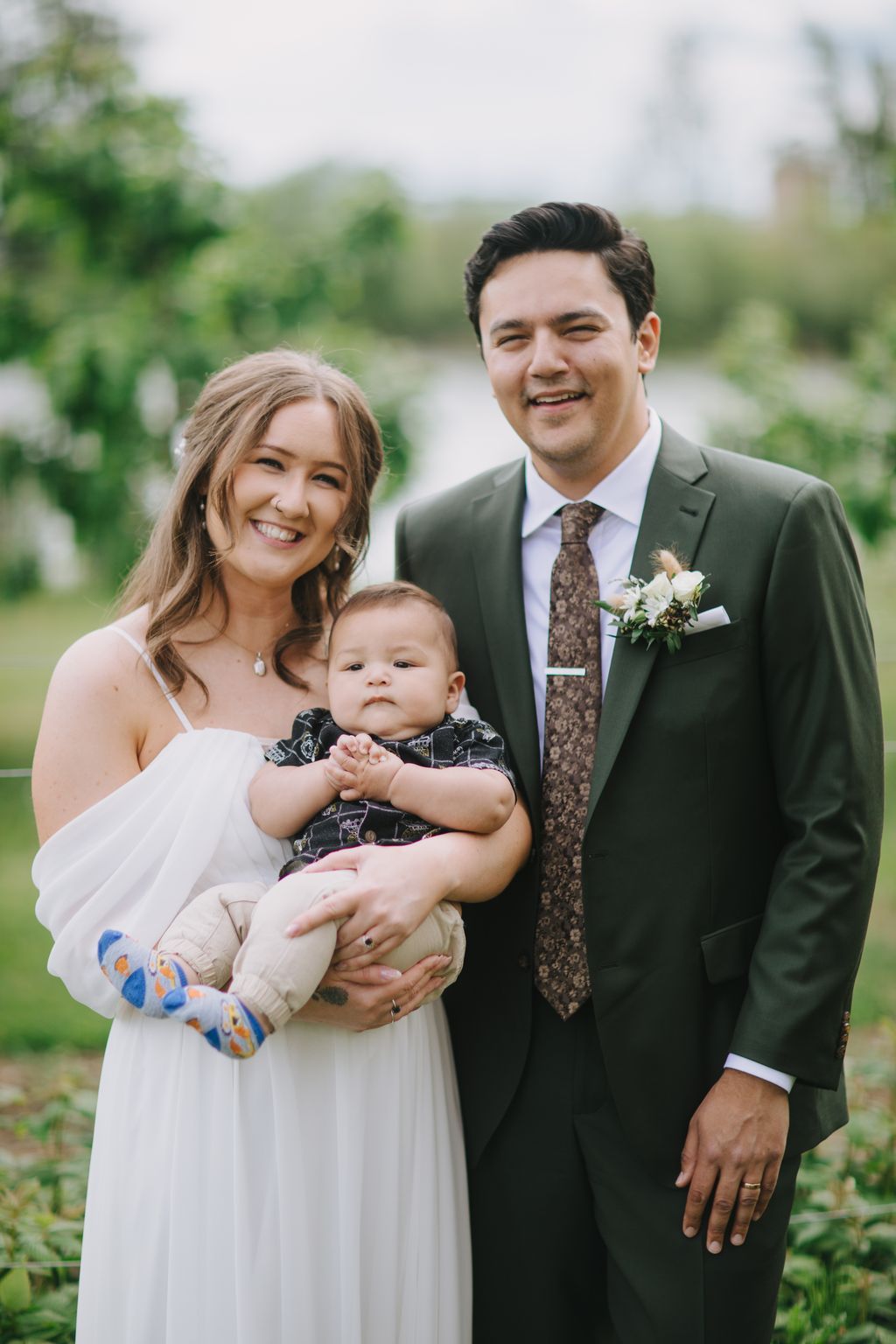 Smiling couple in wedding attire holding their infant son outdoors with blurred green landscape behind