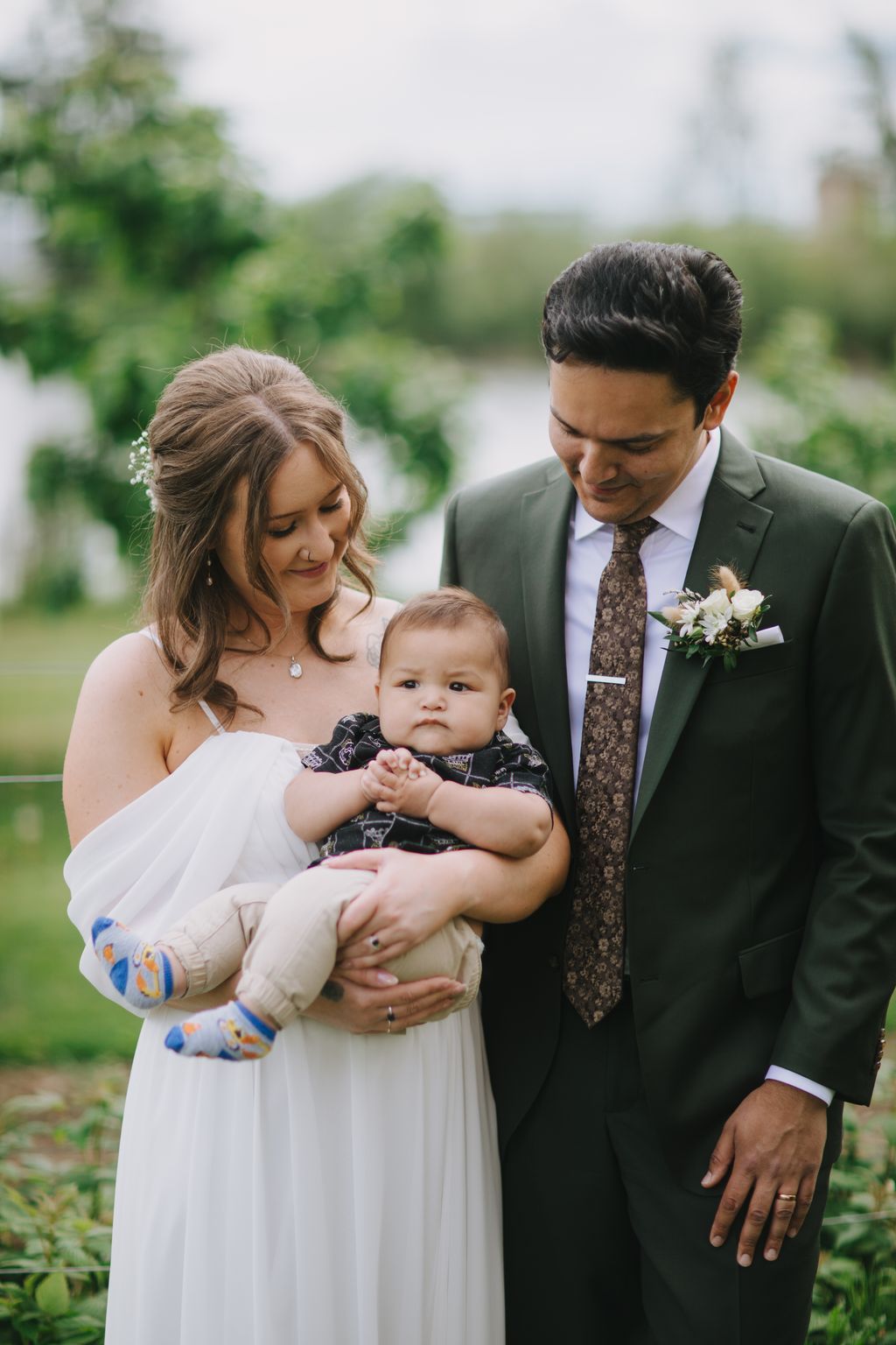 Bride and groom holding their infant son outdoors, smiling down at the child with blurred green landscape behind them