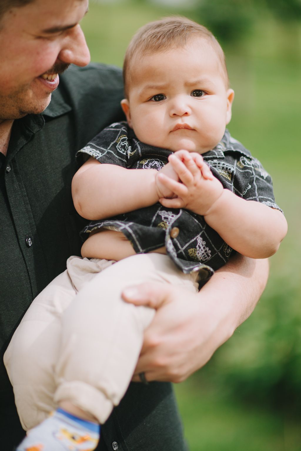 Infant in black patterned outfit held by adult against blurred green outdoor background, soft natural daylight