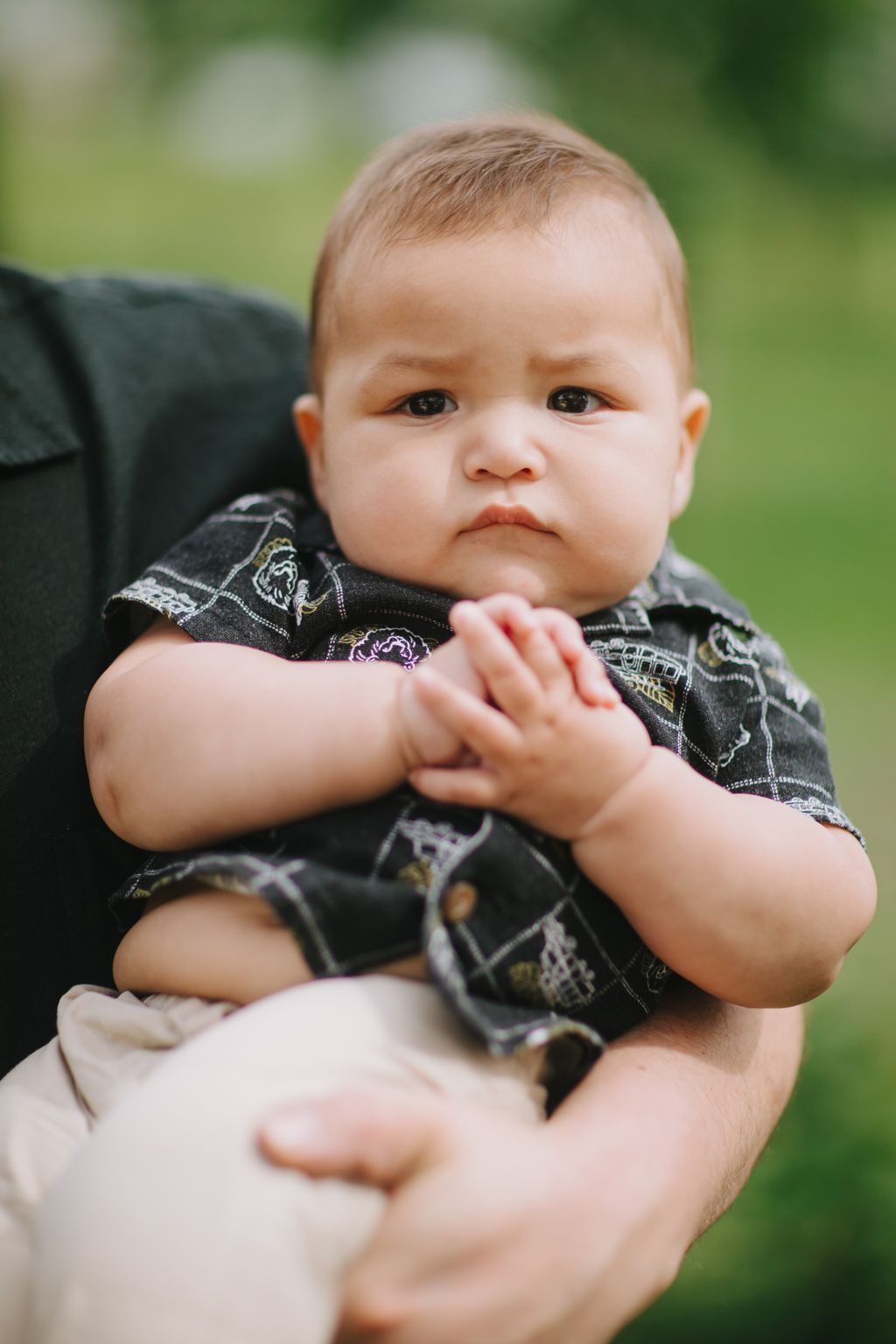 Close-up portrait of a baby in a caregiver''s arms wearing a patterned dark outfit, soft outdoor lighting with blurred green background