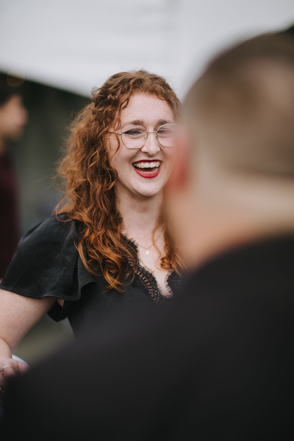 Woman with curly auburn hair and glasses smiling warmly outdoors in natural light