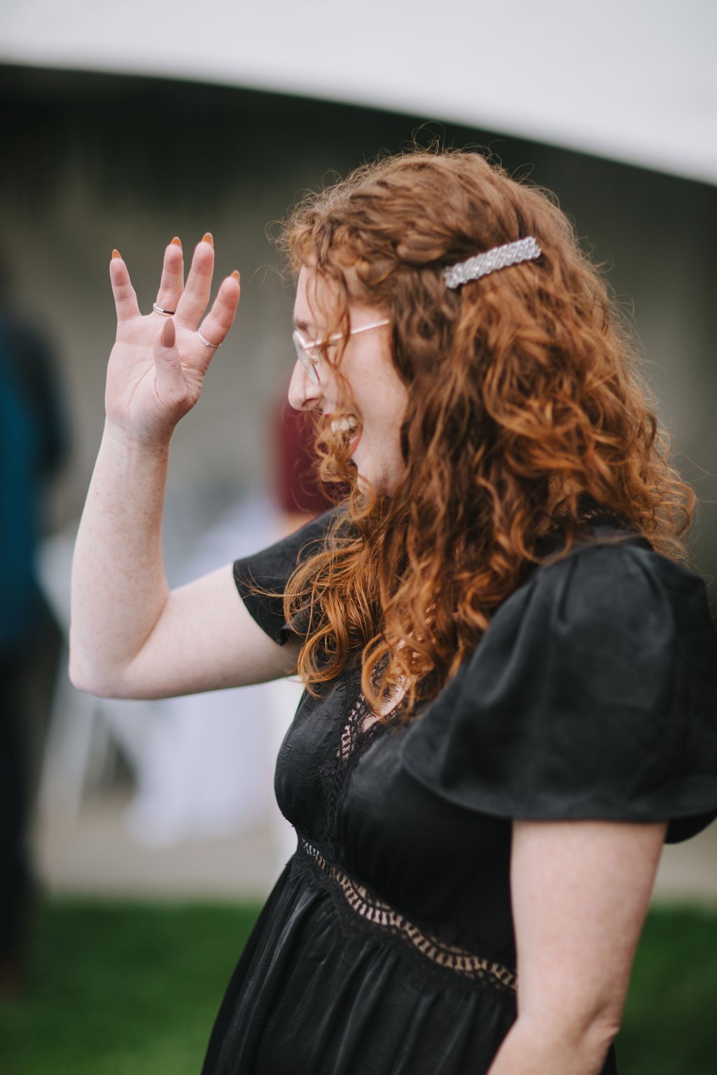 Bride with curly auburn hair and beaded headpiece laughs with hand raised, wearing black lace dress against soft green background